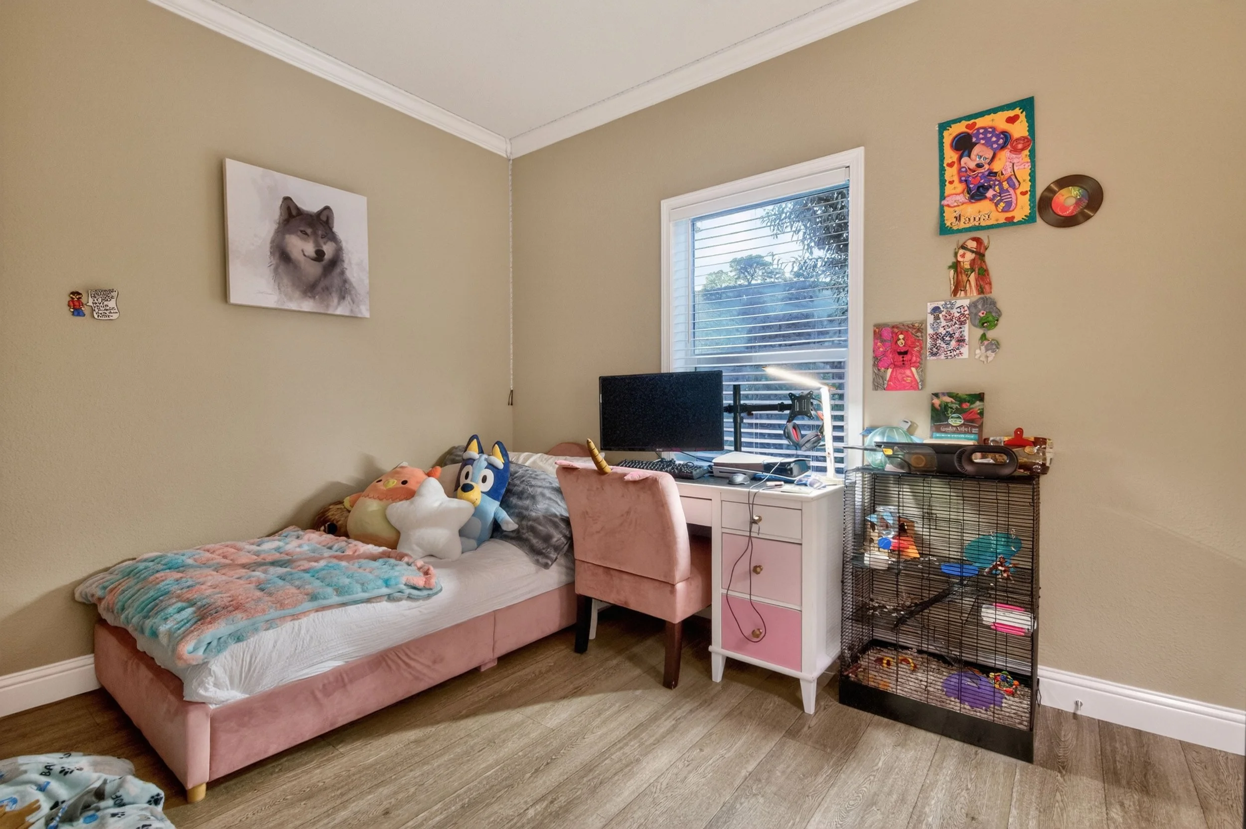 A bedroom with a pink bed frame and plush toys, including a dog, cloud, and animal, on the bed. A window with blinds is above a white desk holding a computer monitor and keyboard. The wall has artwork including a wolf portrait, cartoon characters, an