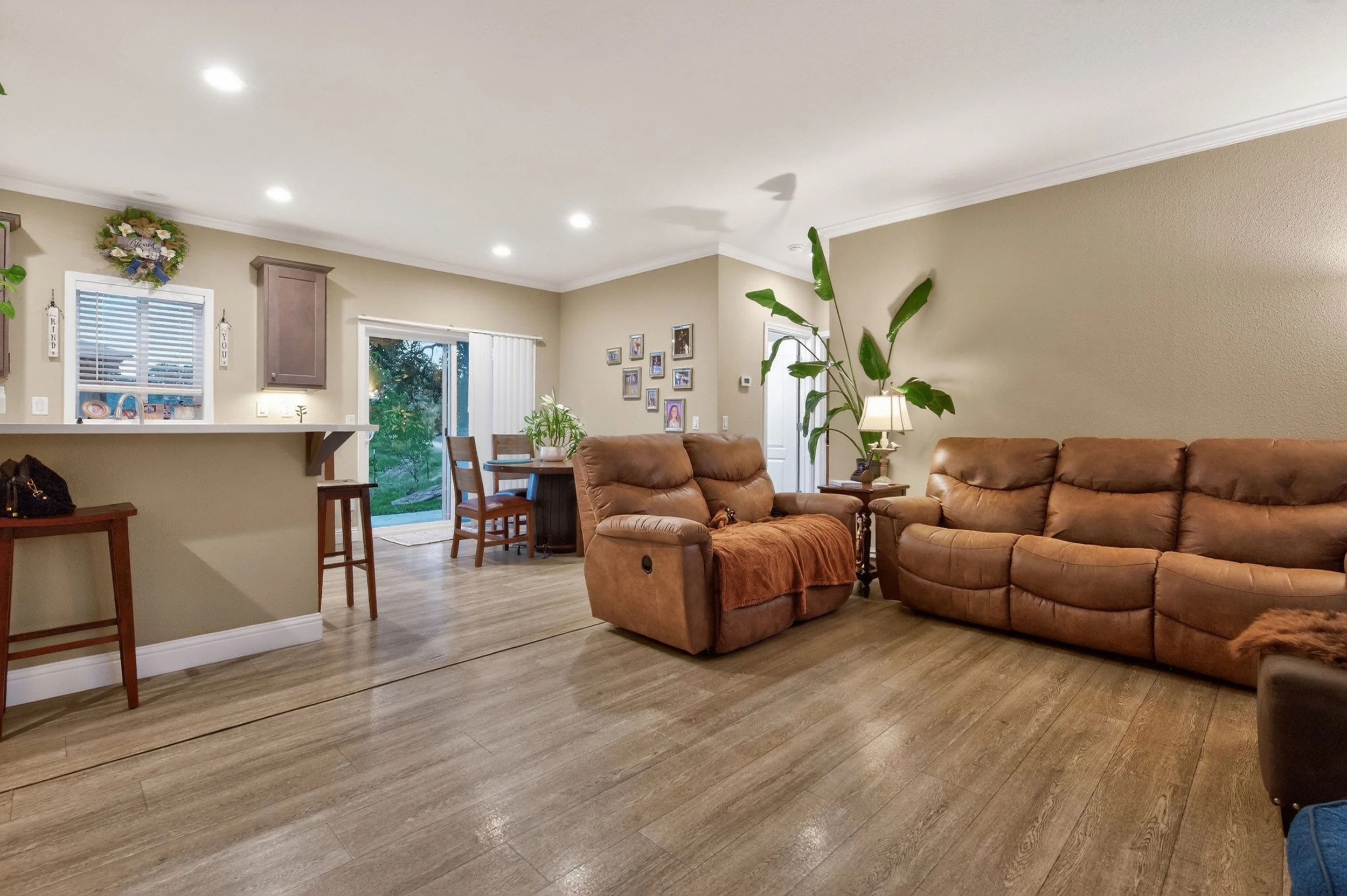 Living room with beige walls, brown leather sofa, green houseplants, framed photos on the wall, wooden dining table with chairs, sliding glass door, and ceiling lights.