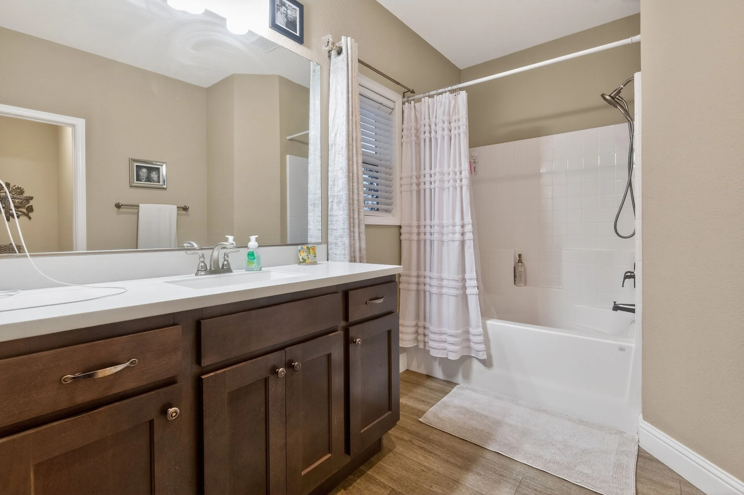 Bathroom with a large mirror above a white countertop vanity with dark wooden cabinets, a towel hanging on the wall, framed photos, a window with blinds and curtains, a white tile bathtub with a shower head, a bottle of shampoo or body wash, and a li