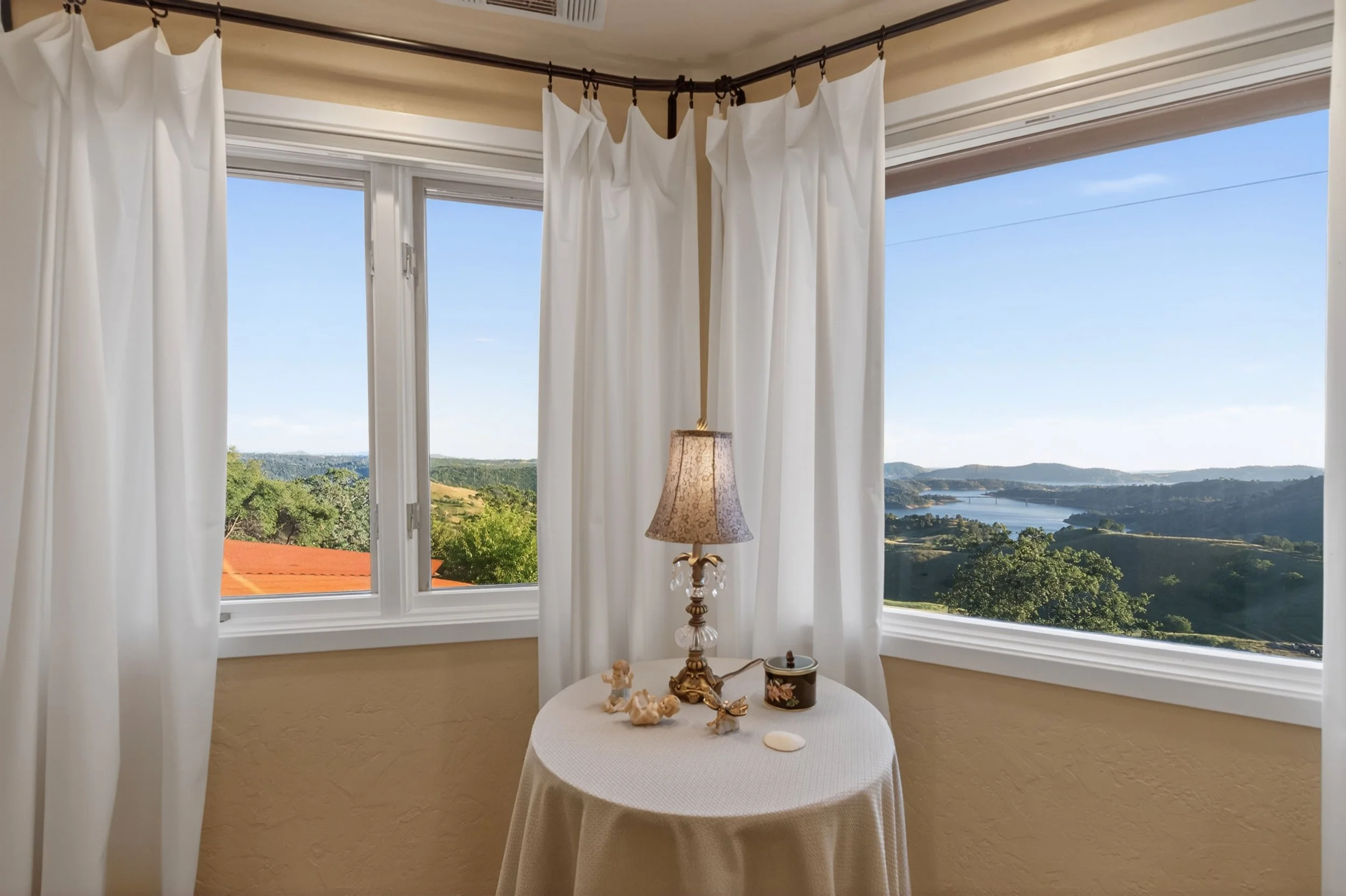 Sunlit living room with large bay window, white curtains, view of green hills and water, small round table with decorative items and a lamp.