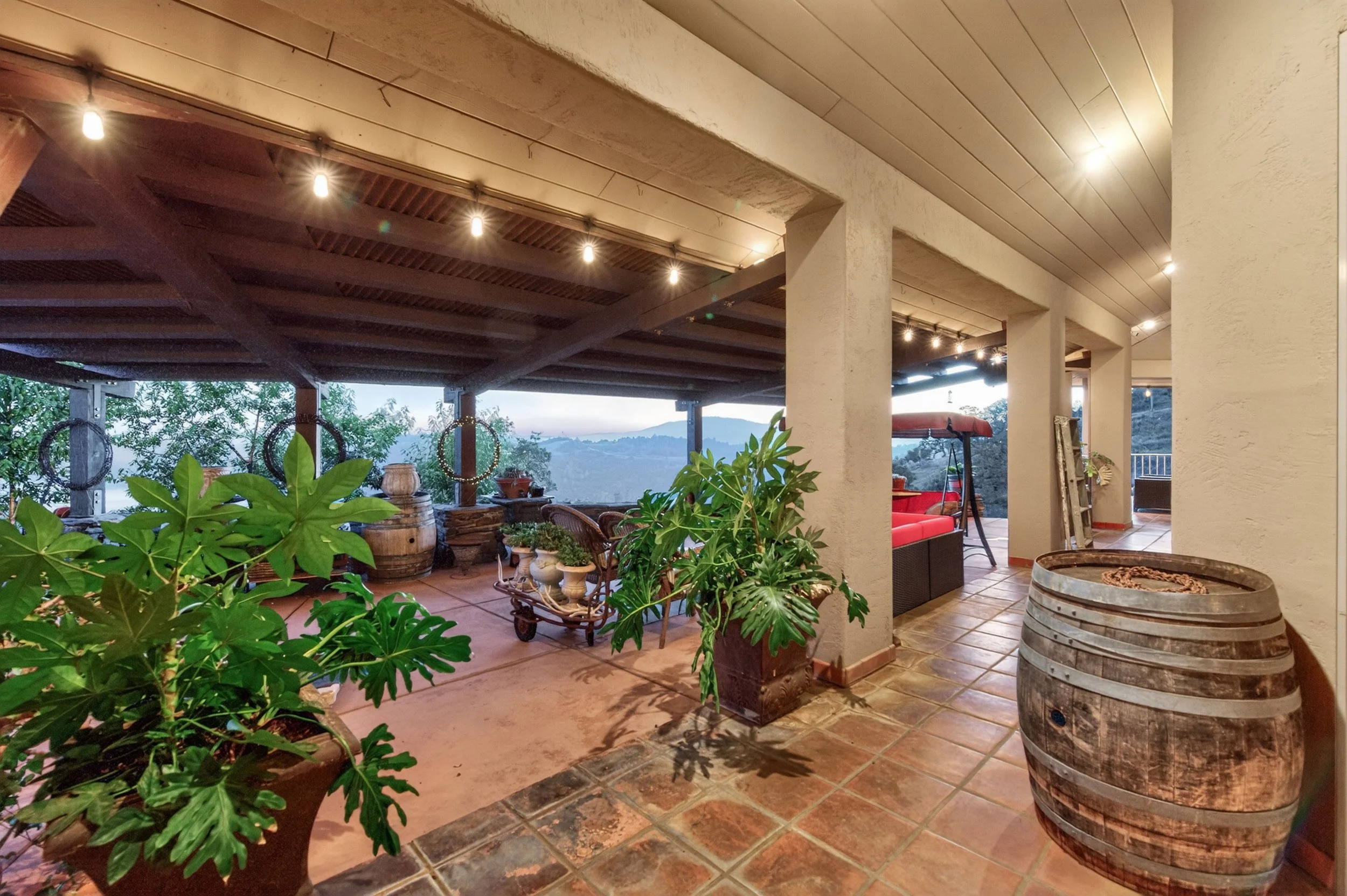 A covered outdoor patio area with plants, wooden barrels, and tile flooring, overlooking a landscape with trees and hills in the distance.