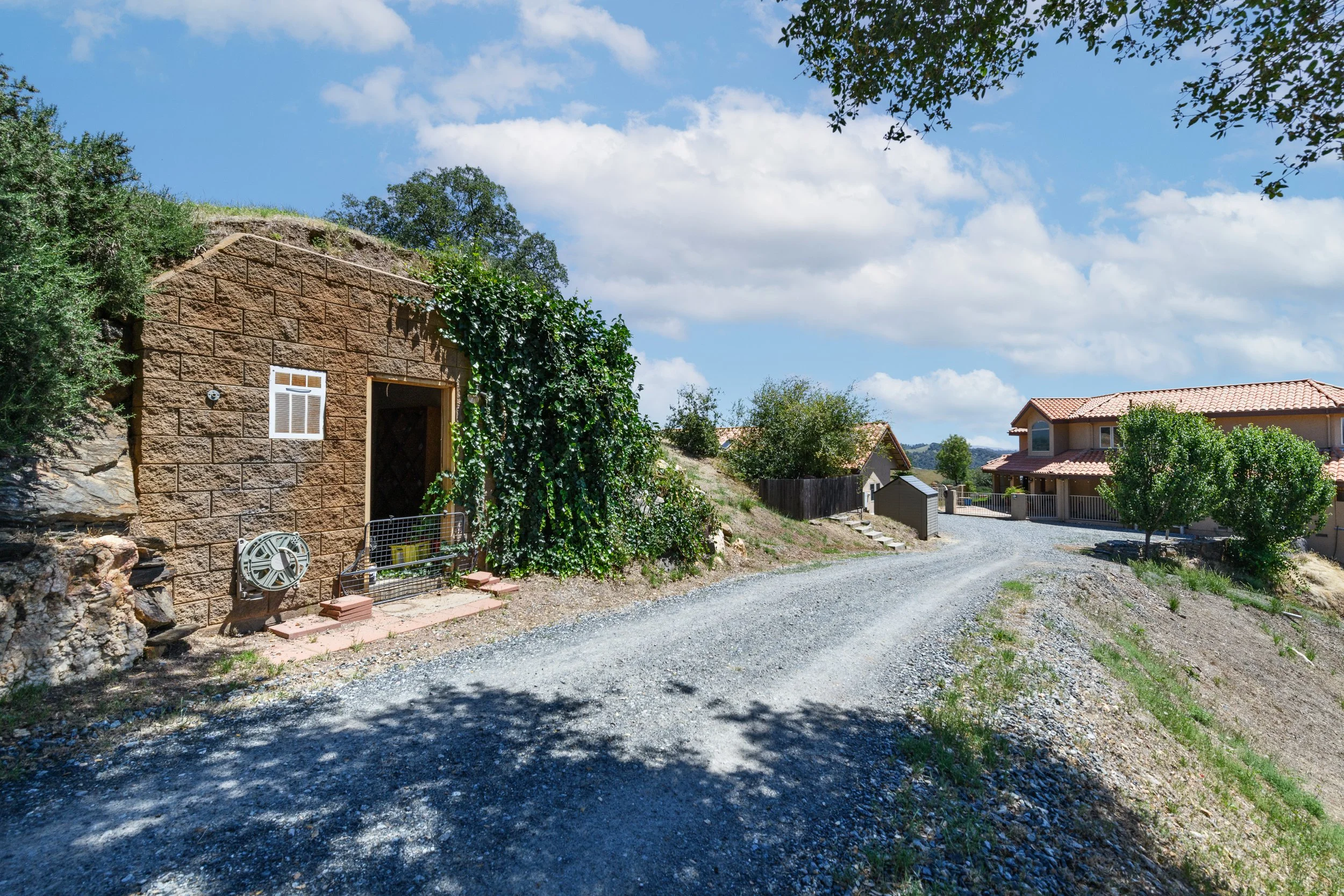 Residential neighborhood with a gravel driveway leading to houses, one with a brick exterior and ivy, under a partly cloudy sky.