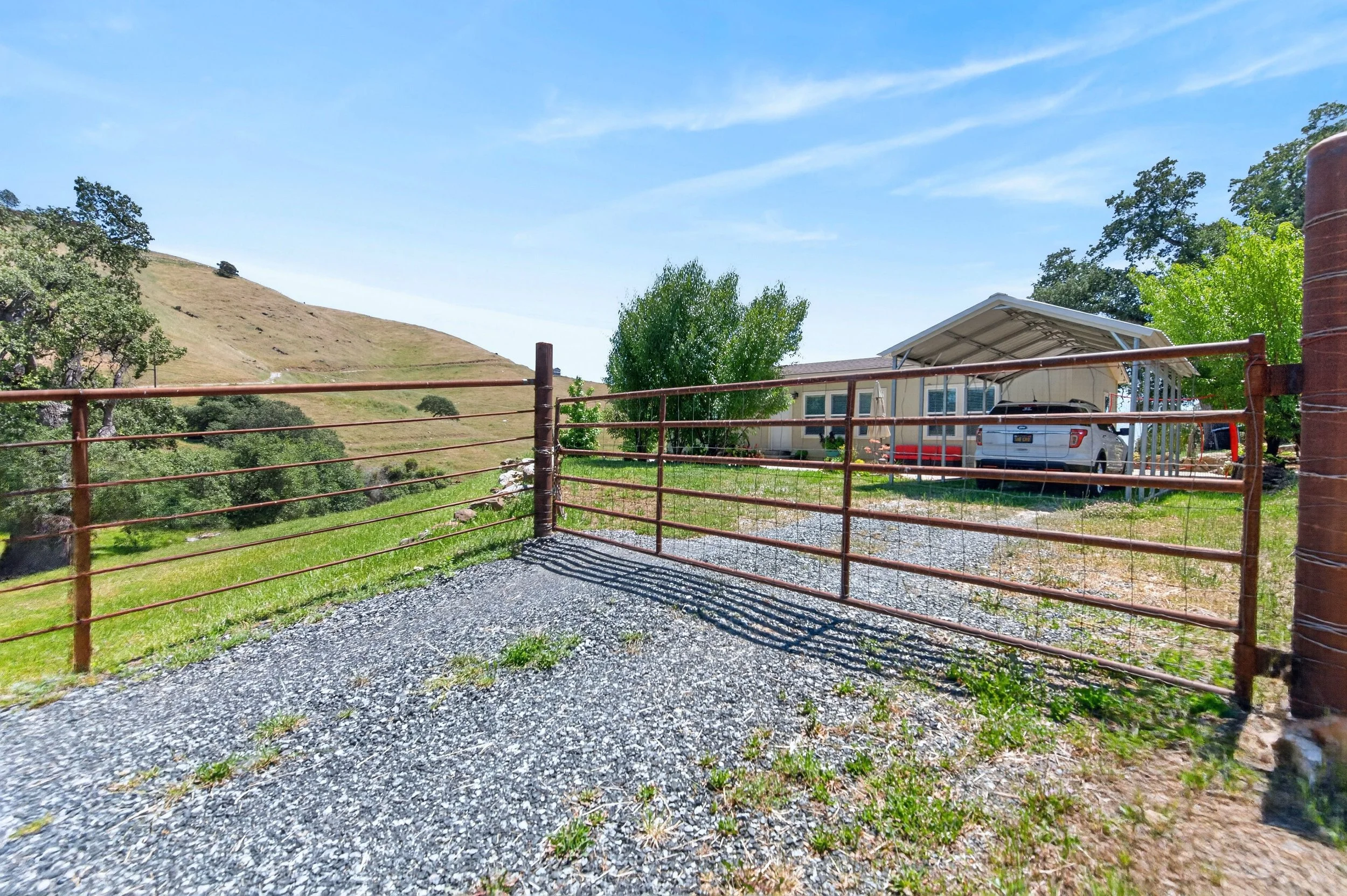 A view of a backyard with a gravel driveway, a metal gate, trees, a house with a carport, and hills in the background under a blue sky.