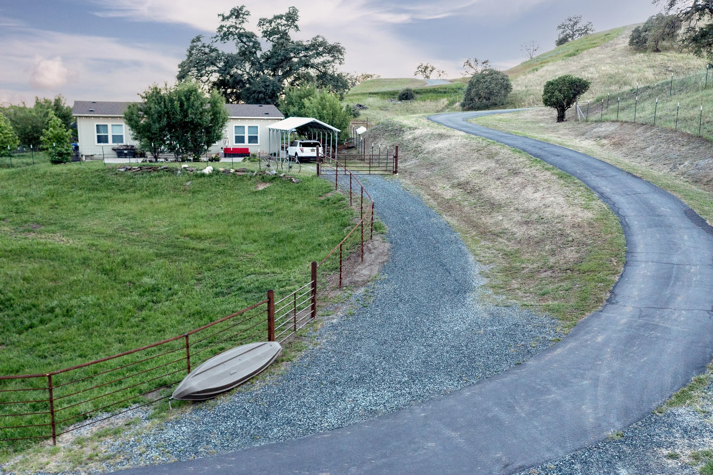 A house with a carport with a white vehicle parked, surrounded by trees and green hills, with a winding driveway and a metal gate.