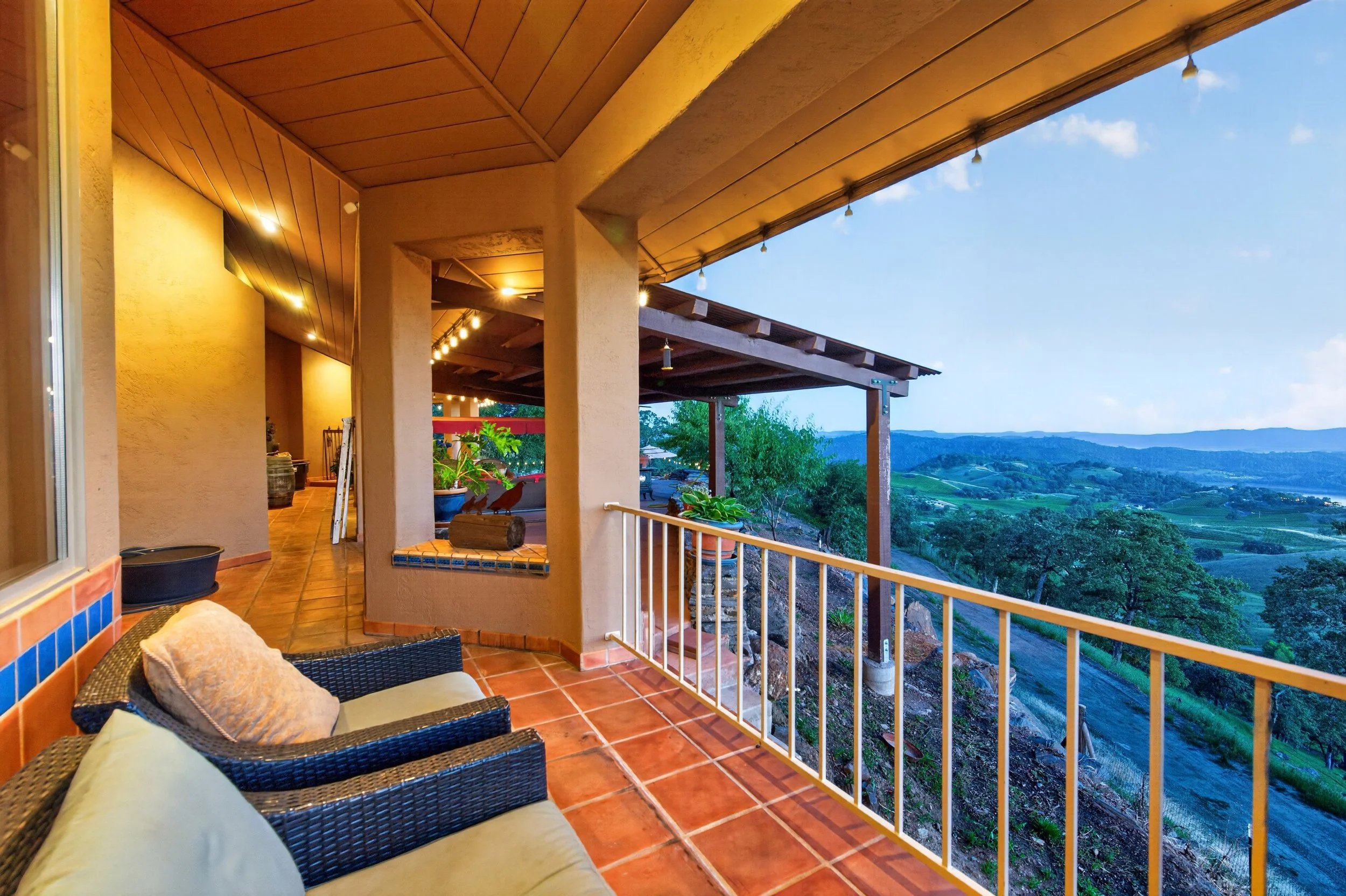 A covered outdoor balcony with terracotta tile flooring, wicker couch, and pillows, overlooking a lush valley with hills and blue sky, lit by string lights at sunset.