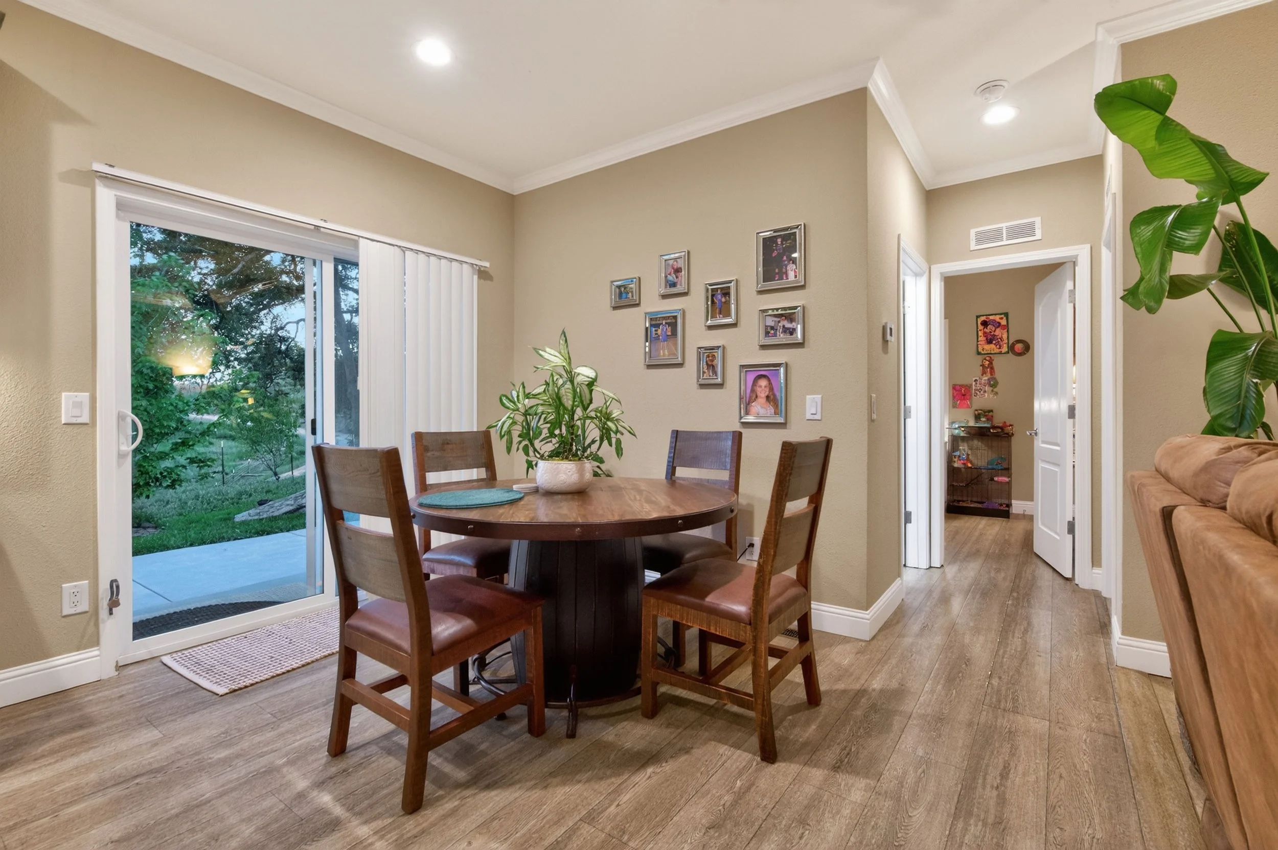 Interior view of a dining area with a round wooden table with chairs, a sliding glass door with vertical blinds leading to a backyard, framed photographs on the wall, and a large green plant.