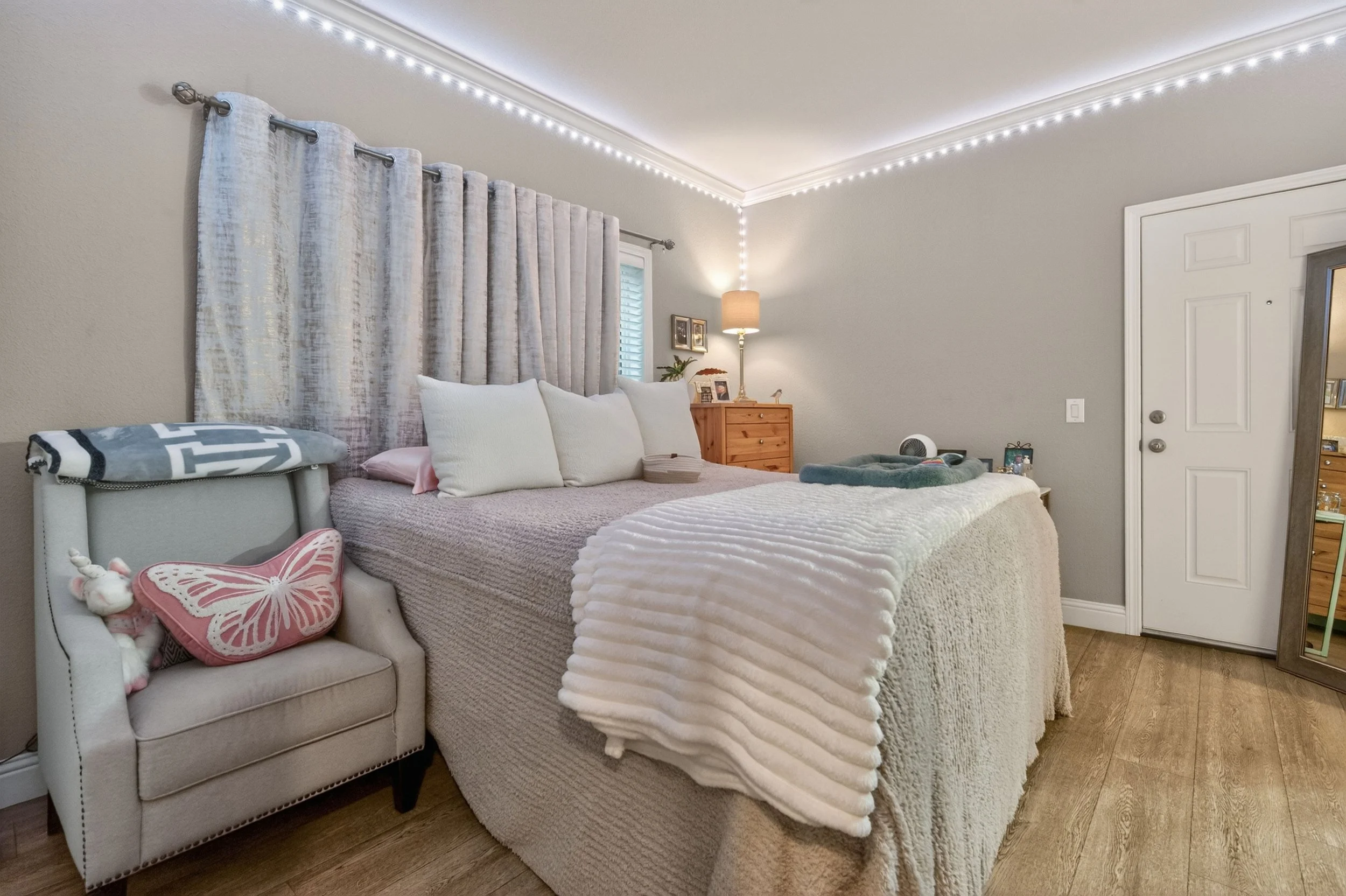 A bedroom with a bed covered in a white blanket, multiple white pillows, and a small side table with a lamp, framed photos, and a plant. A grey armchair with a butterfly pillow and a stuffed unicorn on it is at the foot of the bed. Decorative string 