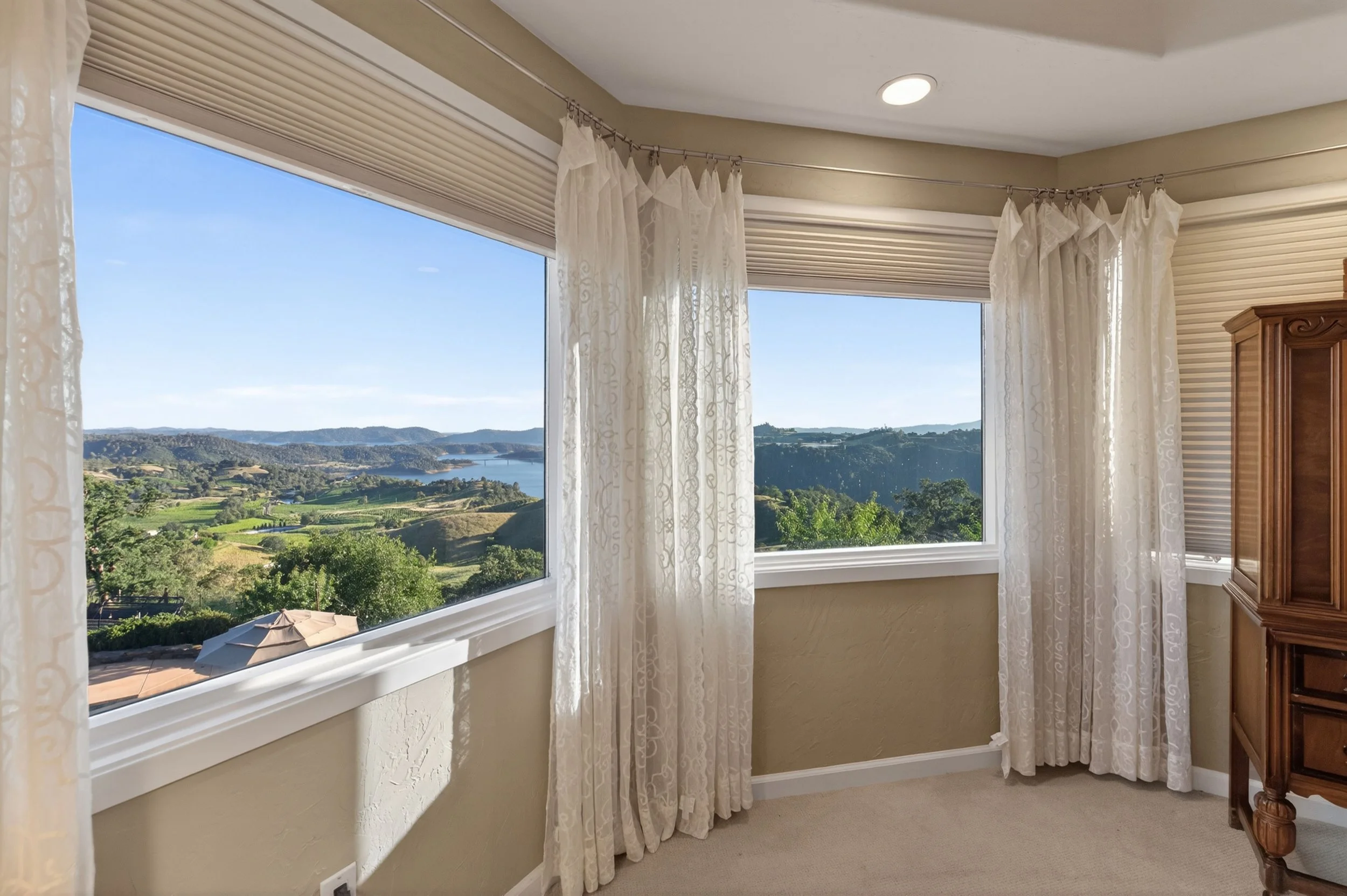 A room with large windows overlooking a scenic landscape of hills, water, and greenery. Light-colored curtains and a wooden cabinet are visible.
