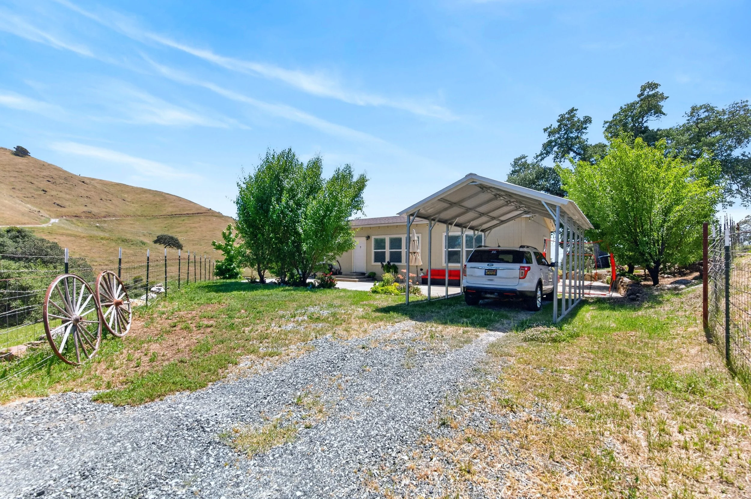 A house with a carport, parked white SUV, green trees, a fence, and a hillside under a blue sky with wispy clouds.