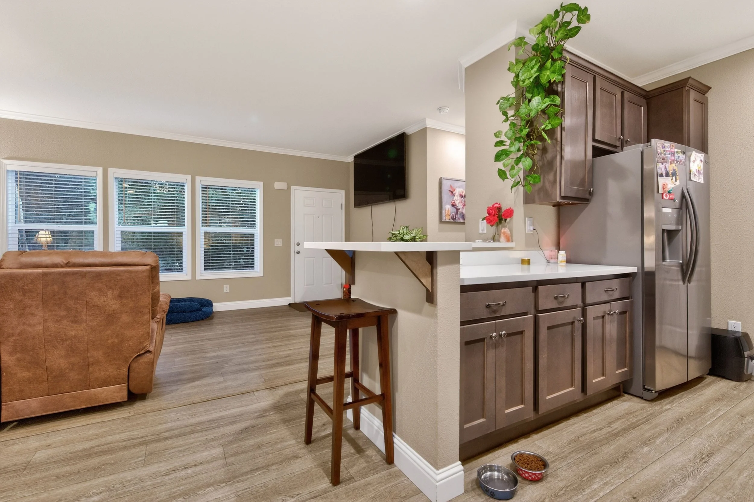 Living room with a window, a brown leather sofa, and a TV mounted on the wall. Partial view of a kitchen with brown cabinets, a stainless steel refrigerator covered with photos and notes, a white counter with a plant and flowers, and pet bowls on the