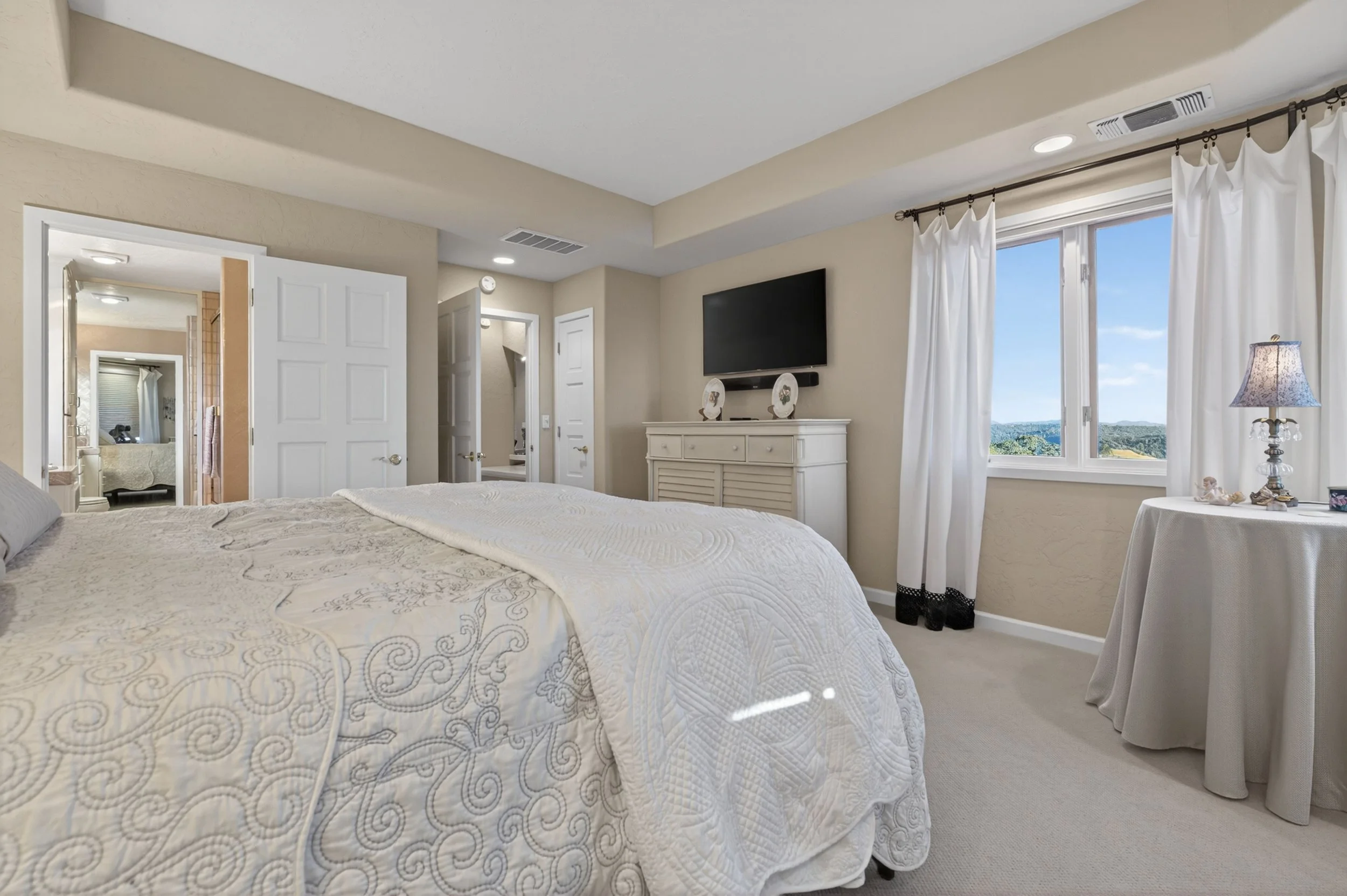 Bedroom with a large bed with a white quilt, a dresser with two decorative plates, a wall-mounted TV, a window with white curtains, and a side table with a lamp.