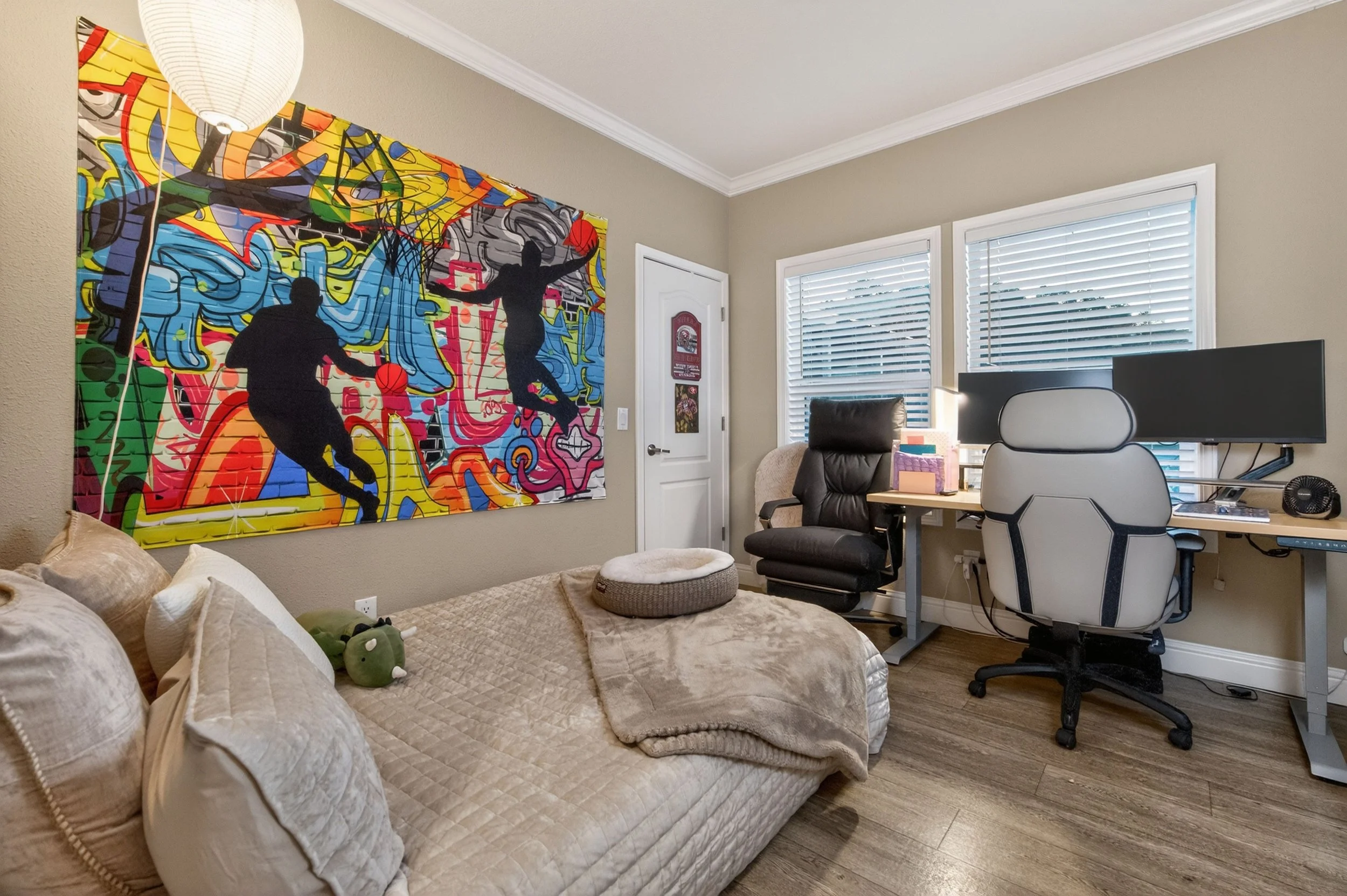 A bedroom featuring a large colorful graffiti-style wall art of basketball players, a bed with beige bedding and pillows, a small plush frog toy, and a workspace with a desk, computer monitor, office chair, and window blinds.
