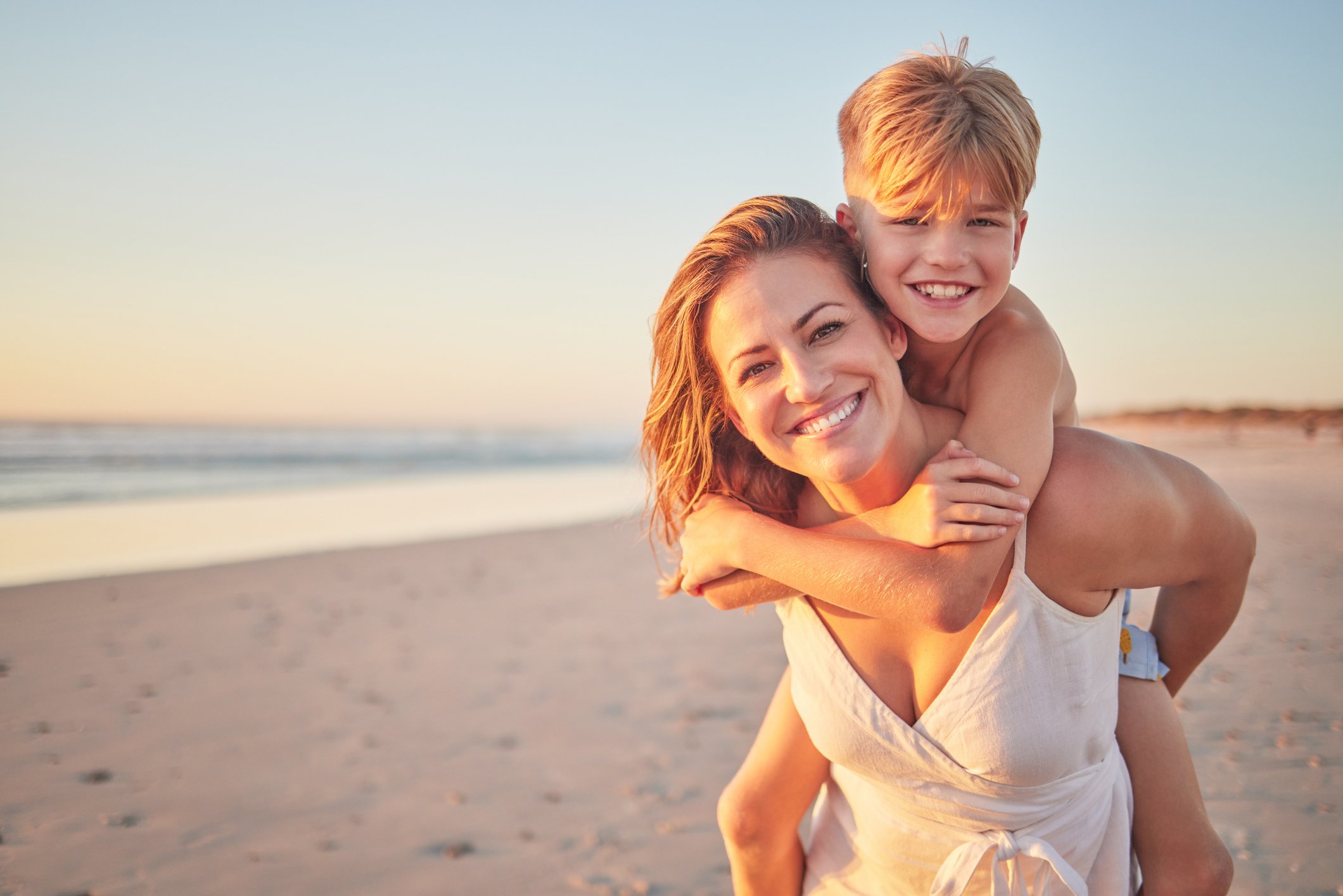 A woman giving a piggyback ride to a young boy on a beach at sunset, both smiling.