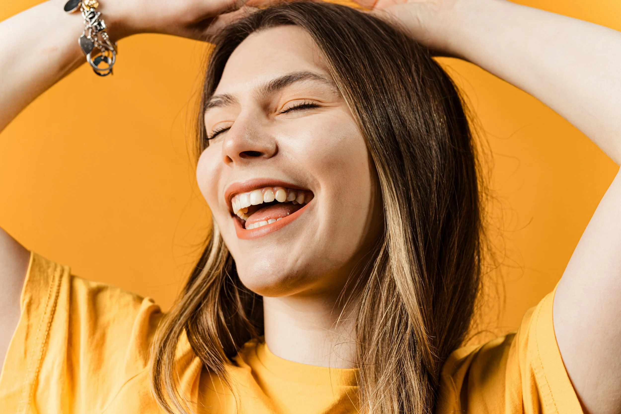 A smiling woman with long brown hair, wearing a yellow top, has her eyes closed and her arms raised above her head against a yellow background.