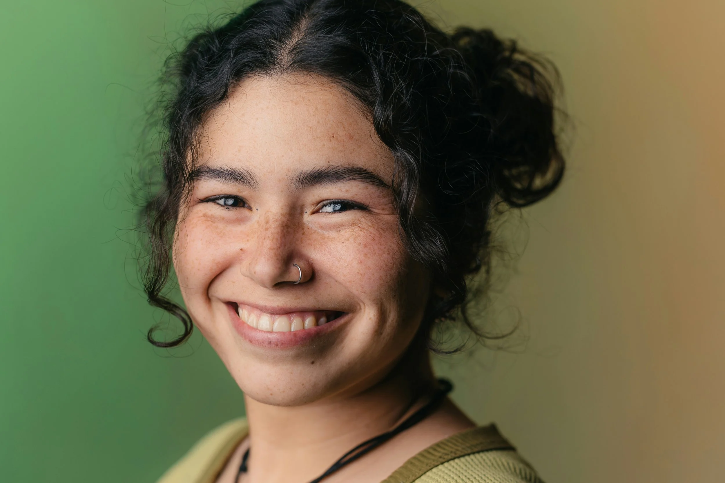 Close-up of a smiling woman with curly dark hair, light skin, and freckles, against a gradient green and yellow background.
