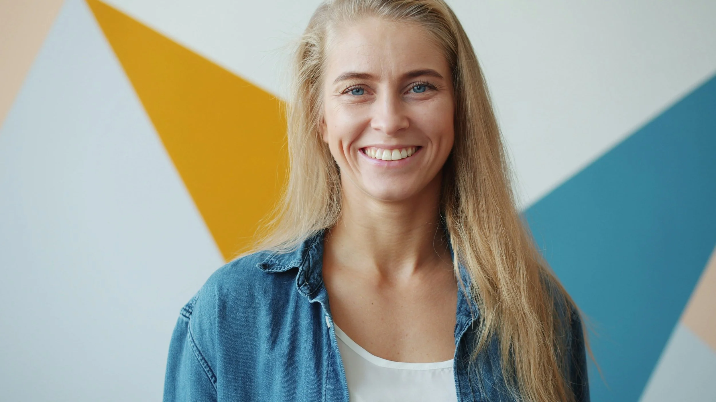 A woman with long blonde hair, blue eyes, and a bright smile, wearing a denim jacket and white top, standing in front of a wall with yellow and blue geometric shapes.