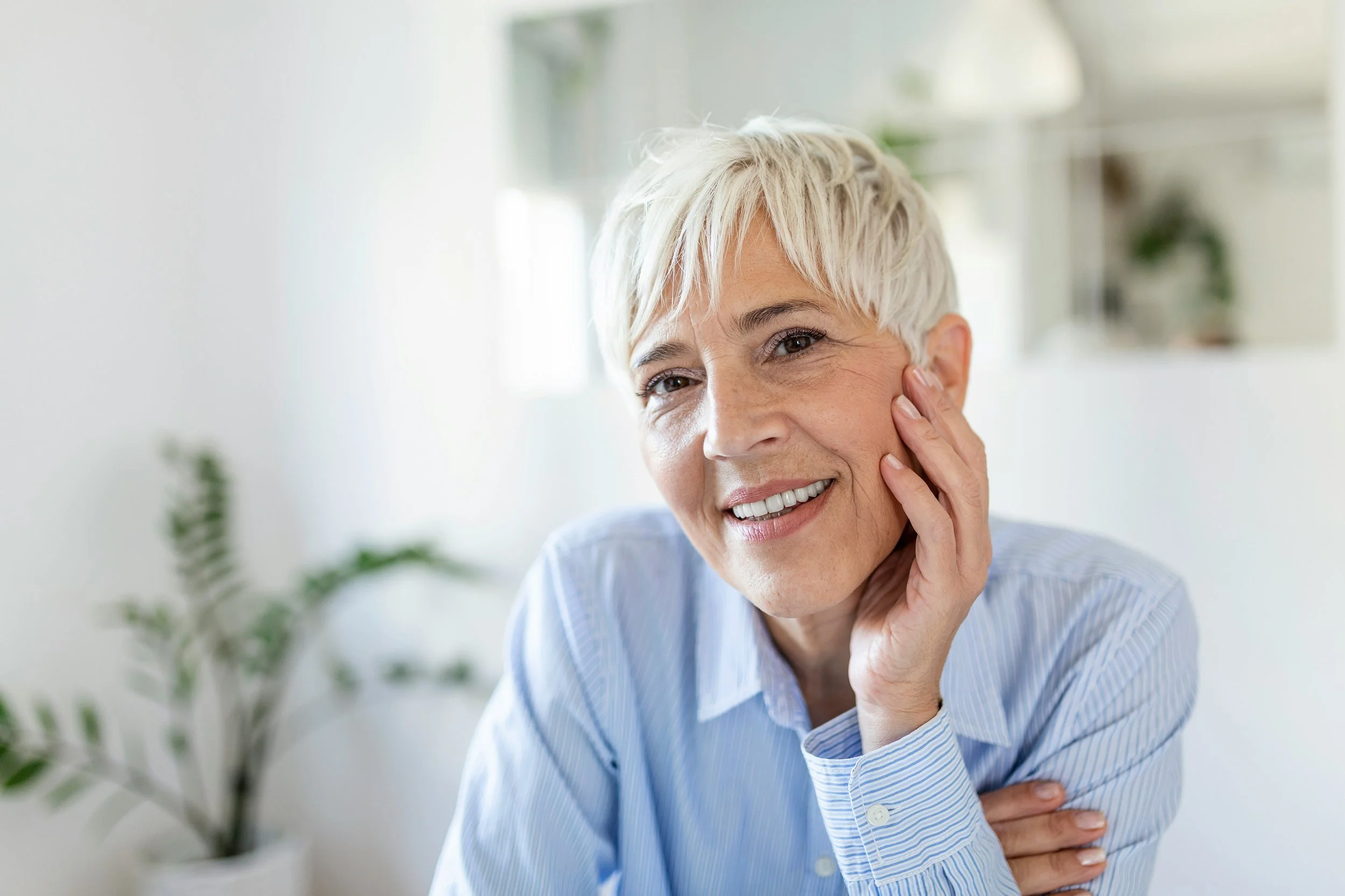 Close-up of a smiling older woman with short white hair, touching her cheek with one hand, wearing a light blue striped shirt.