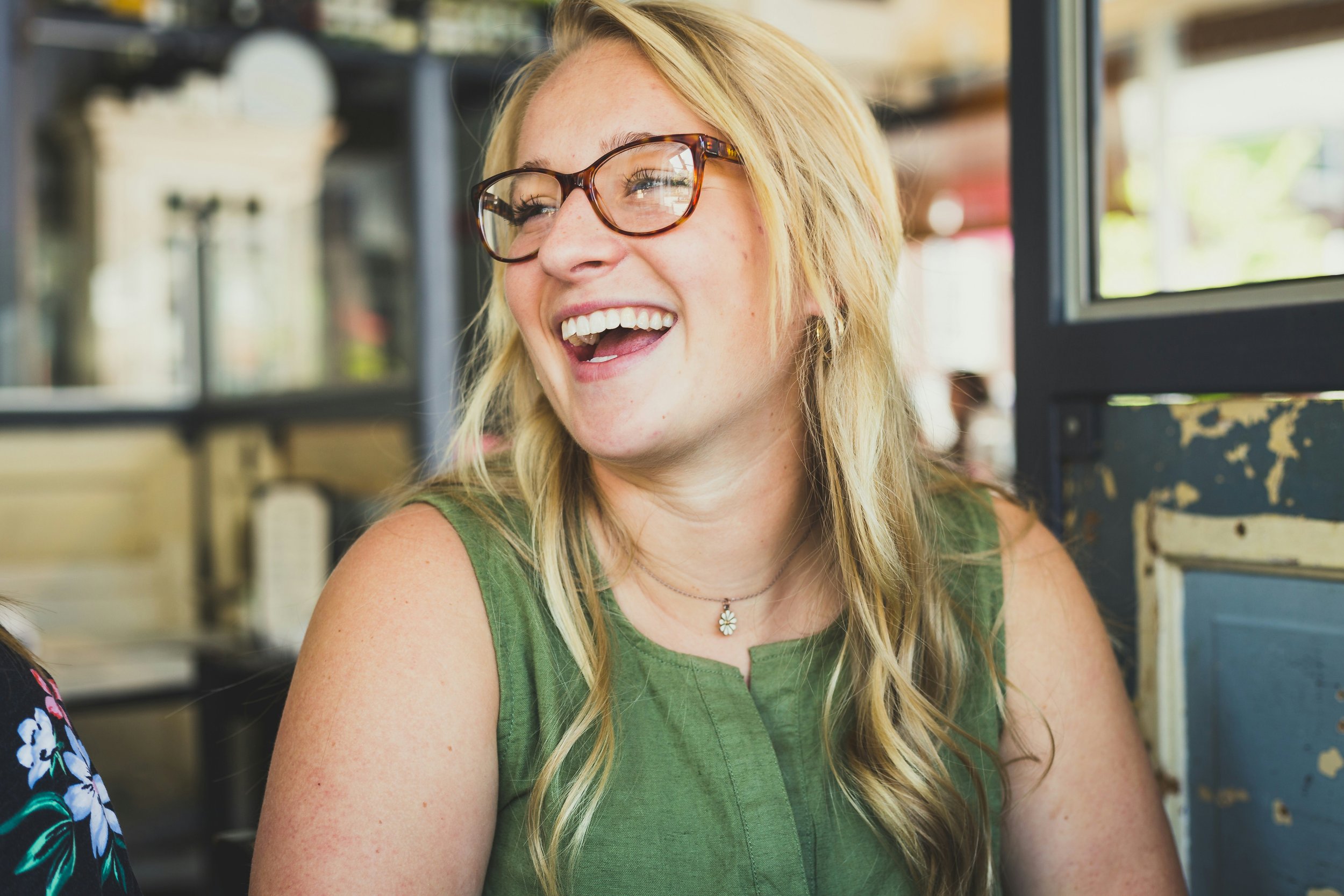 A woman with blond hair and glasses, smiling and looking to the side, in an outdoor or cafe setting.