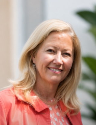 A smiling woman with blonde hair, wearing a coral-colored jacket and earrings, standing outdoors near greenery.