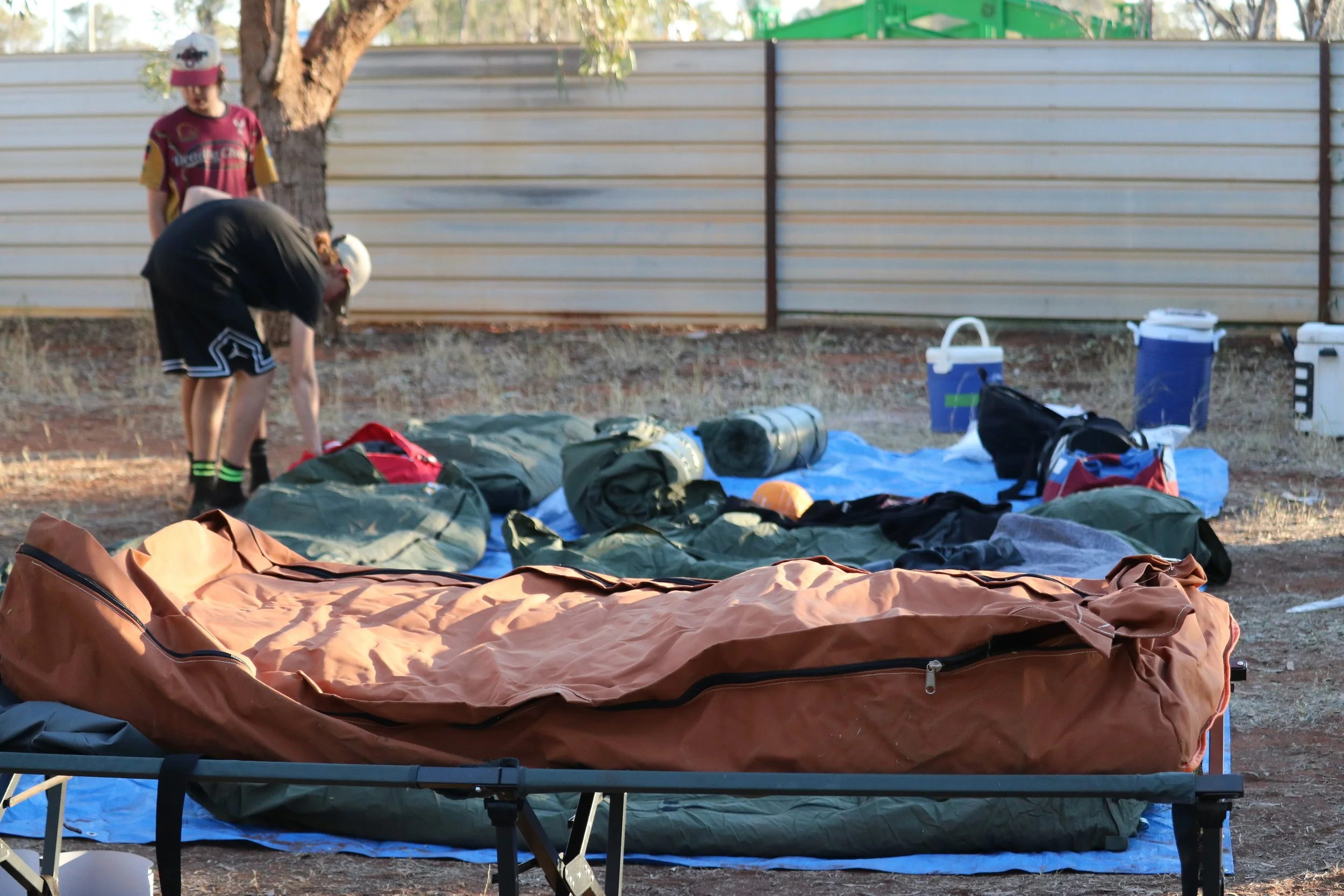 Outdoor scene with sleeping area set up on a table, including a brown tent and sleeping gear, with two children in baseball caps organizing items nearby. Background features a wooden fence and some trees.