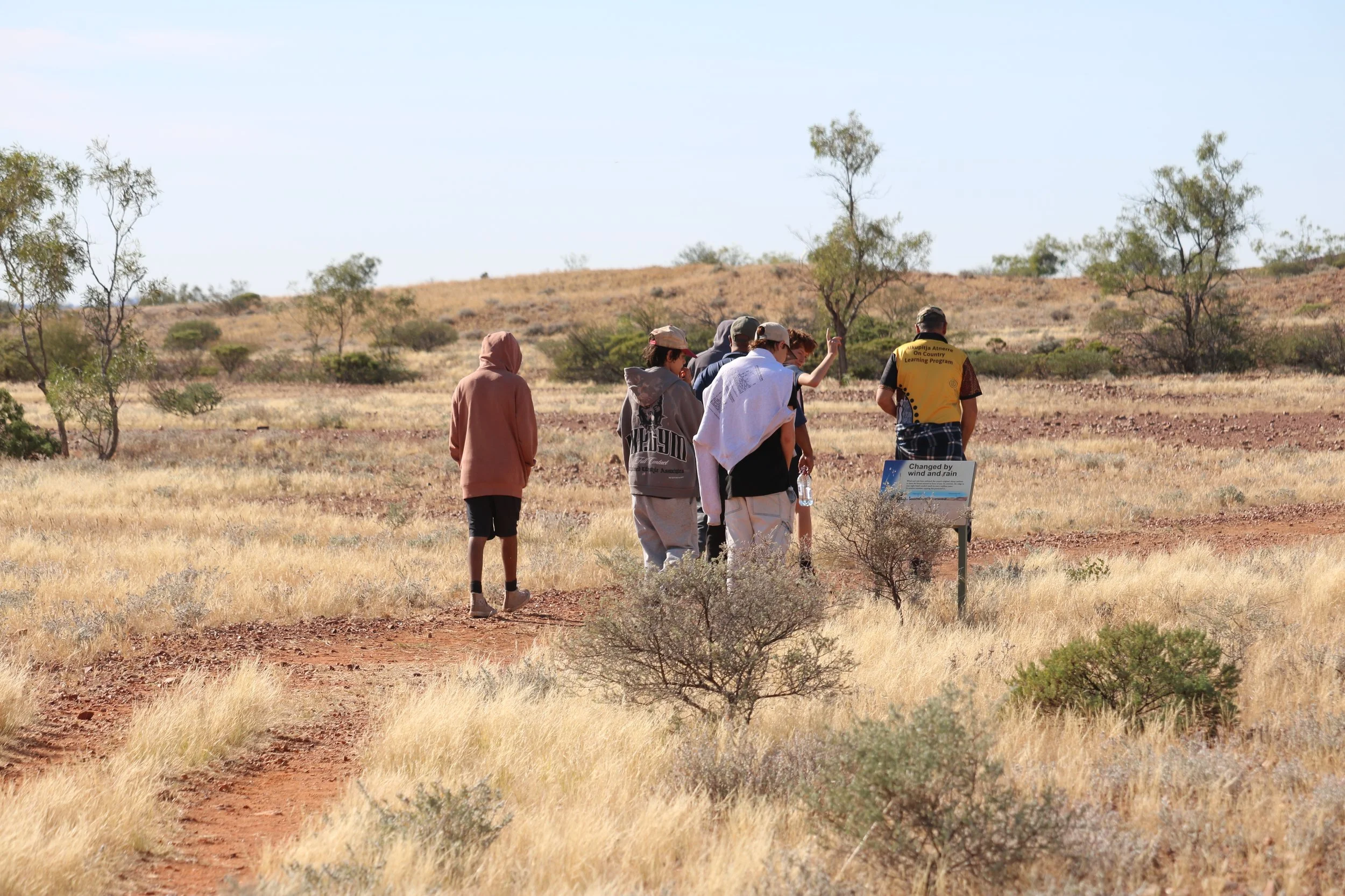 A group of people, including children and adults, walking through a dry, grassy, open landscape with sparse trees, on a sunny day. Some are wearing hats and casual outdoor clothing.