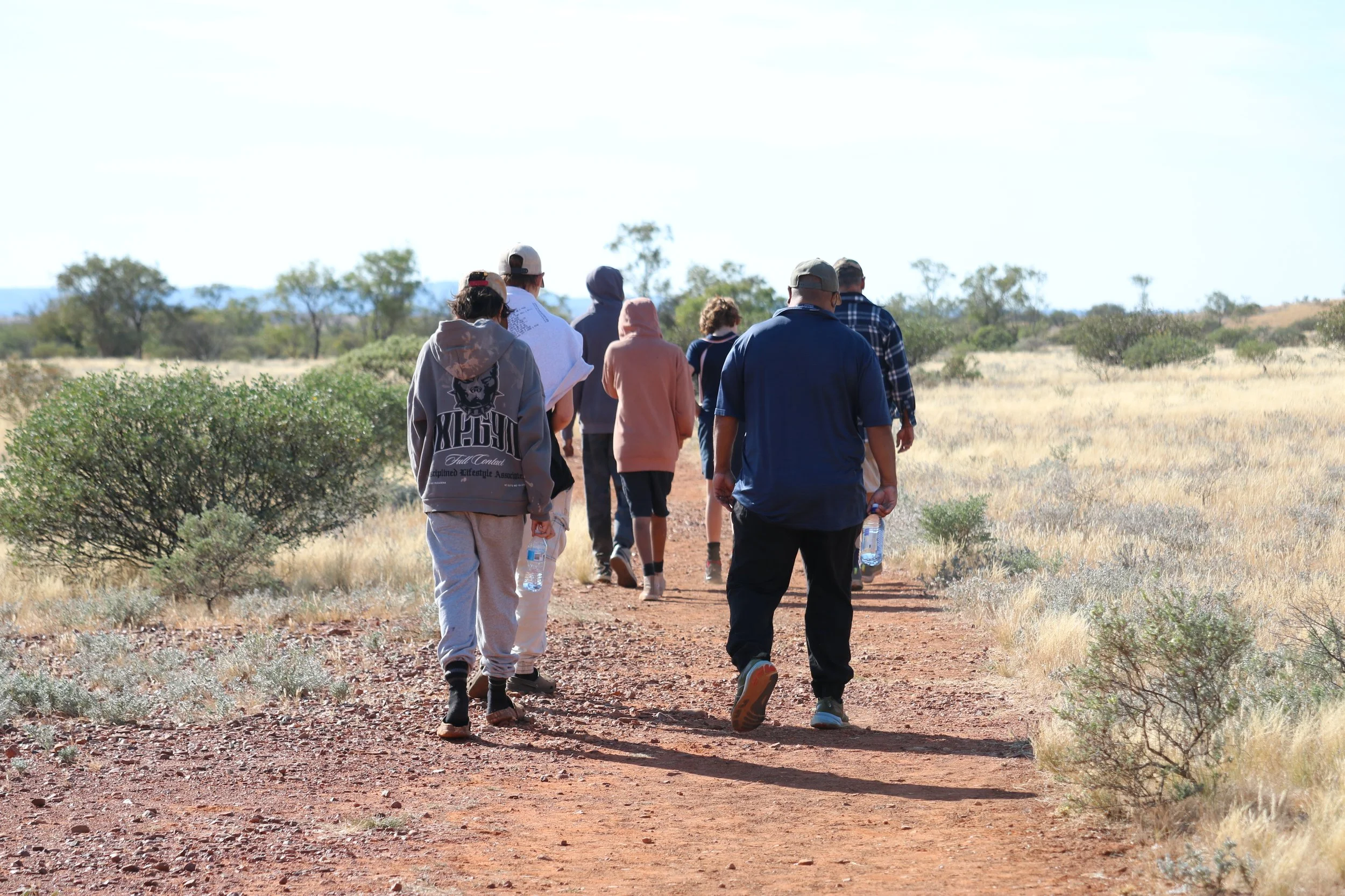 A group of people hiking on a dirt trail in a grassy, semi-arid landscape with sparse trees under a partly cloudy sky.