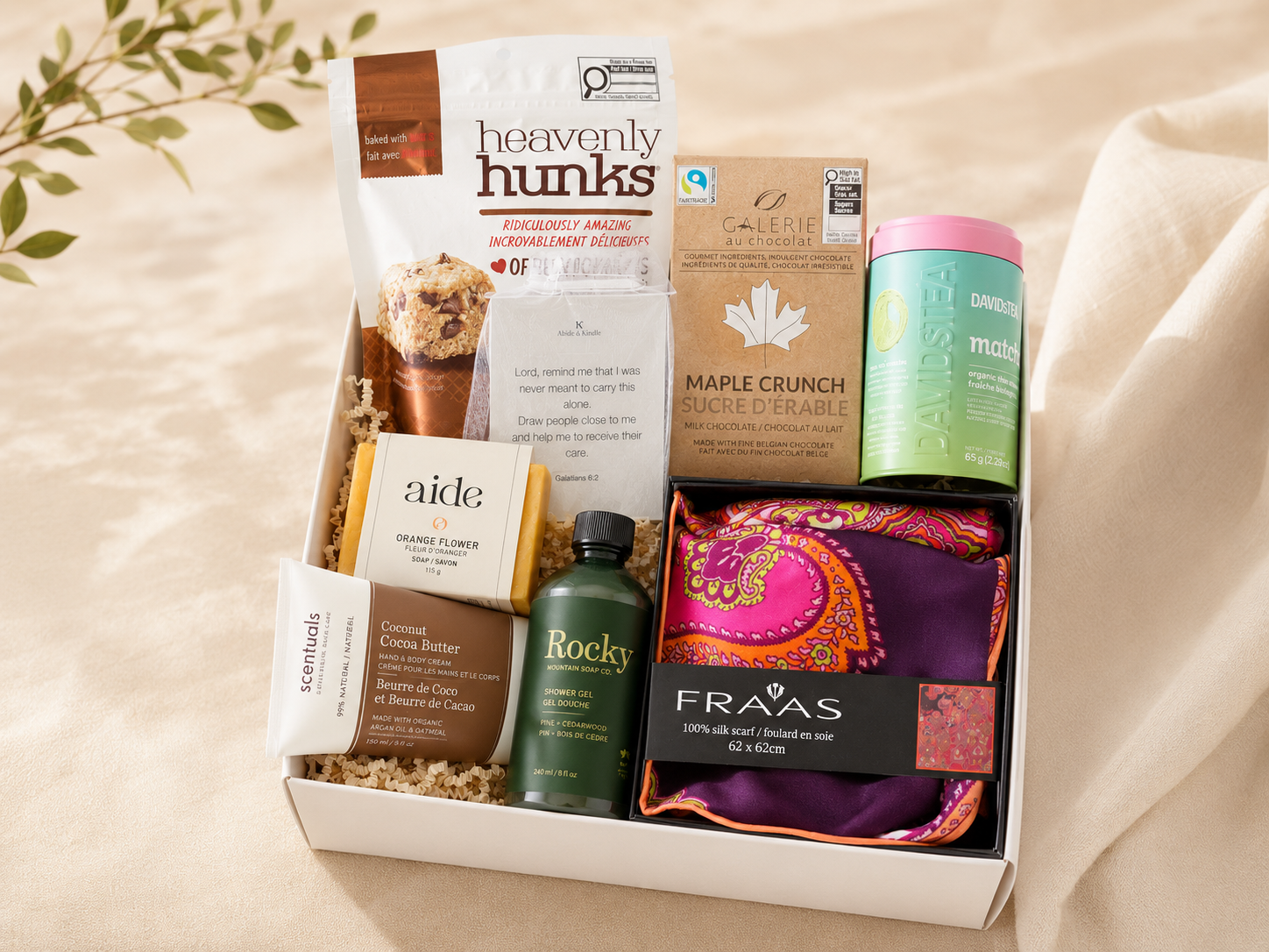 A gift box containing various items including a bag of heavenly hunks cookies, a box of Galerie au chocolat Maple Crunch, a Dambustea matcha organic tea container, a bar of Aide orange flower soap, a small jar of coconut cocoa butter, a Rocky shower gel, a folded colorful silk scarf, and a card with a religious quote and plant and branch in the background.