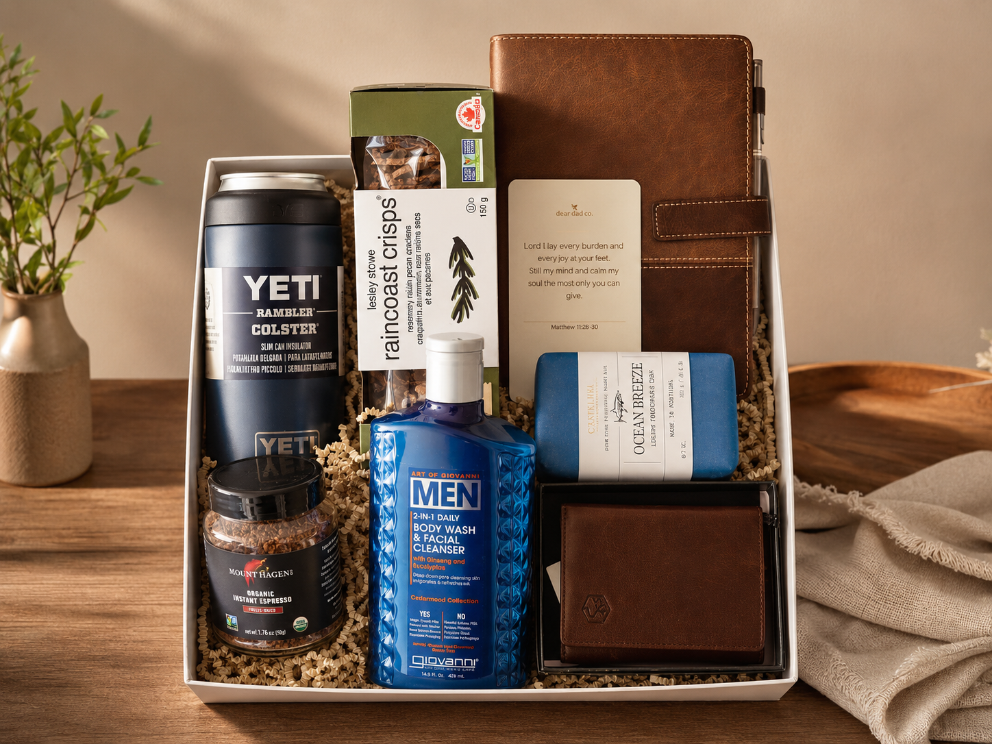 A gift box containing a YETI Rambler tumbler, a box of Rainforest Crisps, a card, a blue bottle of Art of Giovanni Men's body wash, a blue soap bar, a brown leather wallet, a small jar of Mount Hagen instant espresso, a brown leather planner, and a small brown leather box, displayed on a wooden table with a potted plant.