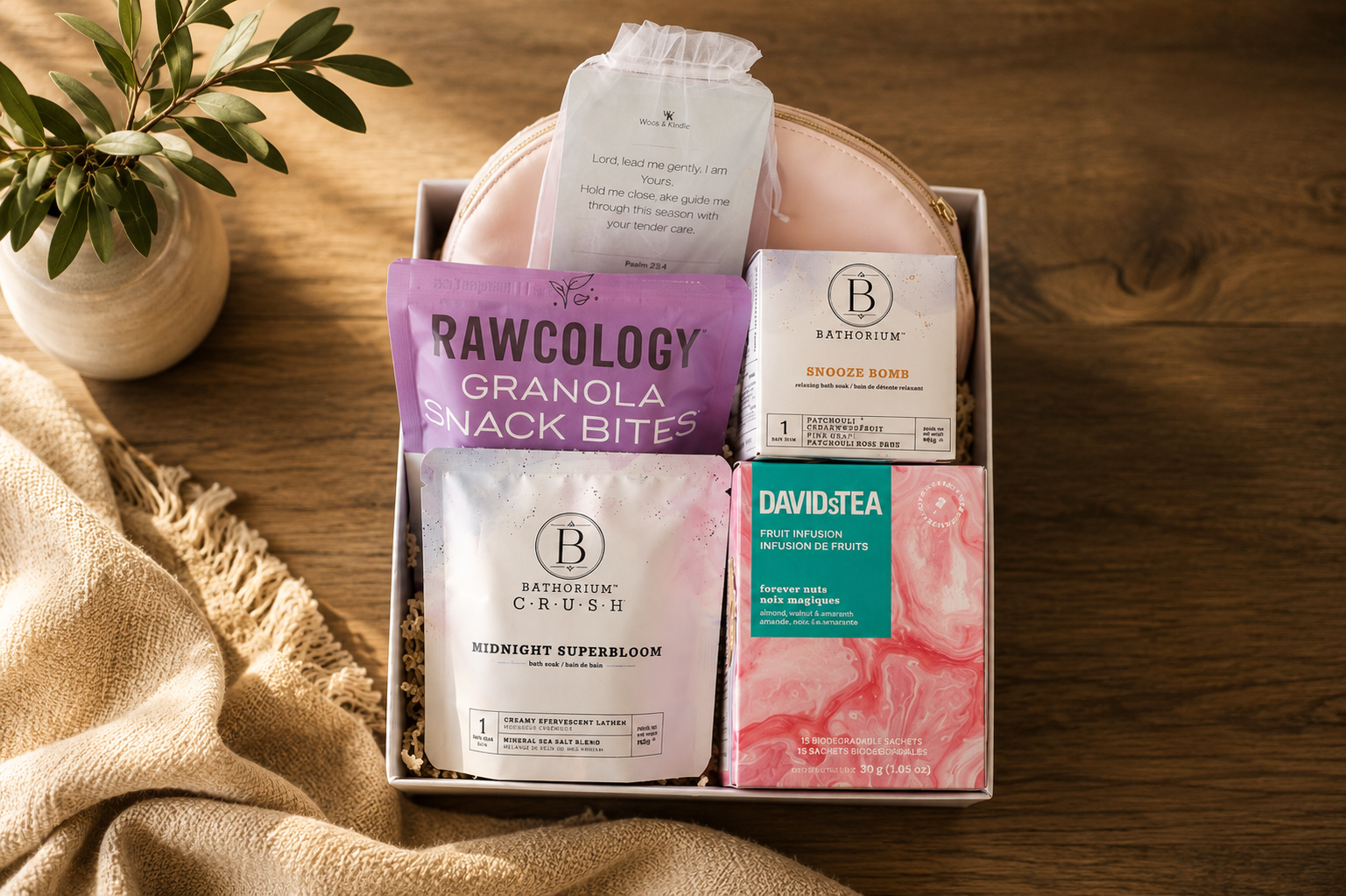A gift box containing bath and wellness products, including a packet of Rawcology granola snack bites, Bathorium Midnight Superbloom bath soak, Bathorium Snooze Bomb relaxing bath soak, David's Tea fruit infusion, and a small book with a prayer.