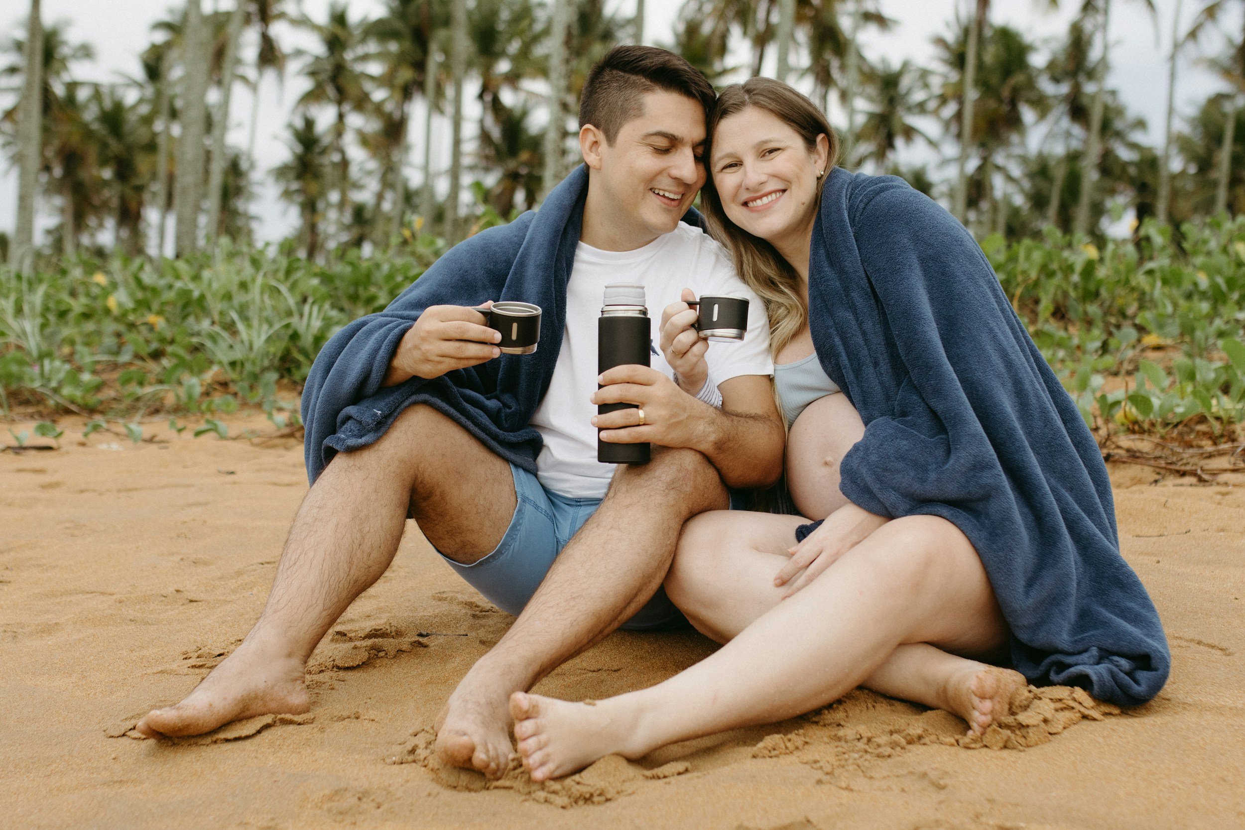 Una pareja se sienta en la playa, envueltos en mantas, compartiendo una taza y una botella térmica, sonriendo y disfrutando del momento juntos.