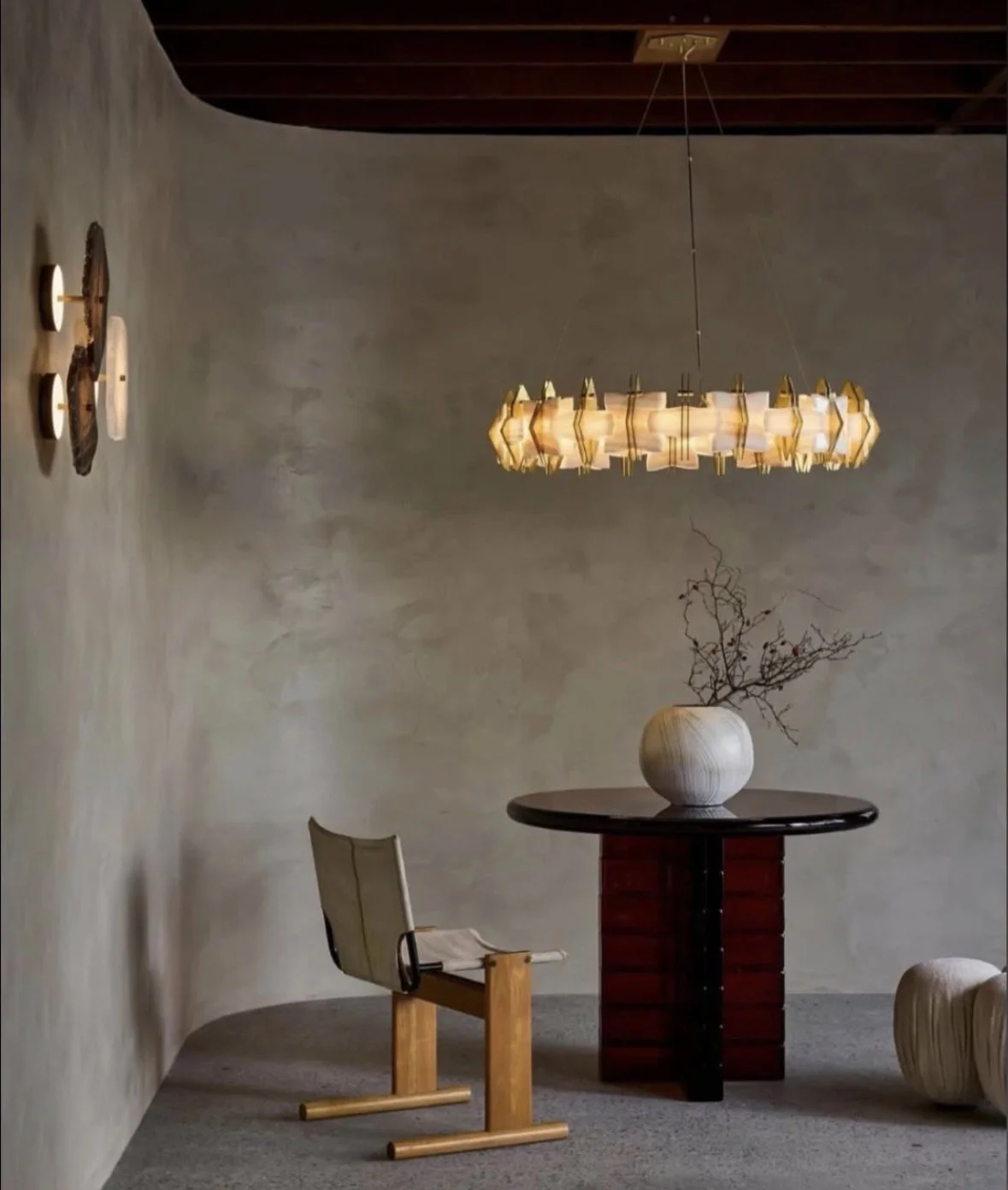 A minimalist interior with a round wooden table, a modern beige chair, a ceramic vase with branches, and a wooden floor lamp, illuminated by a contemporary chandelier.