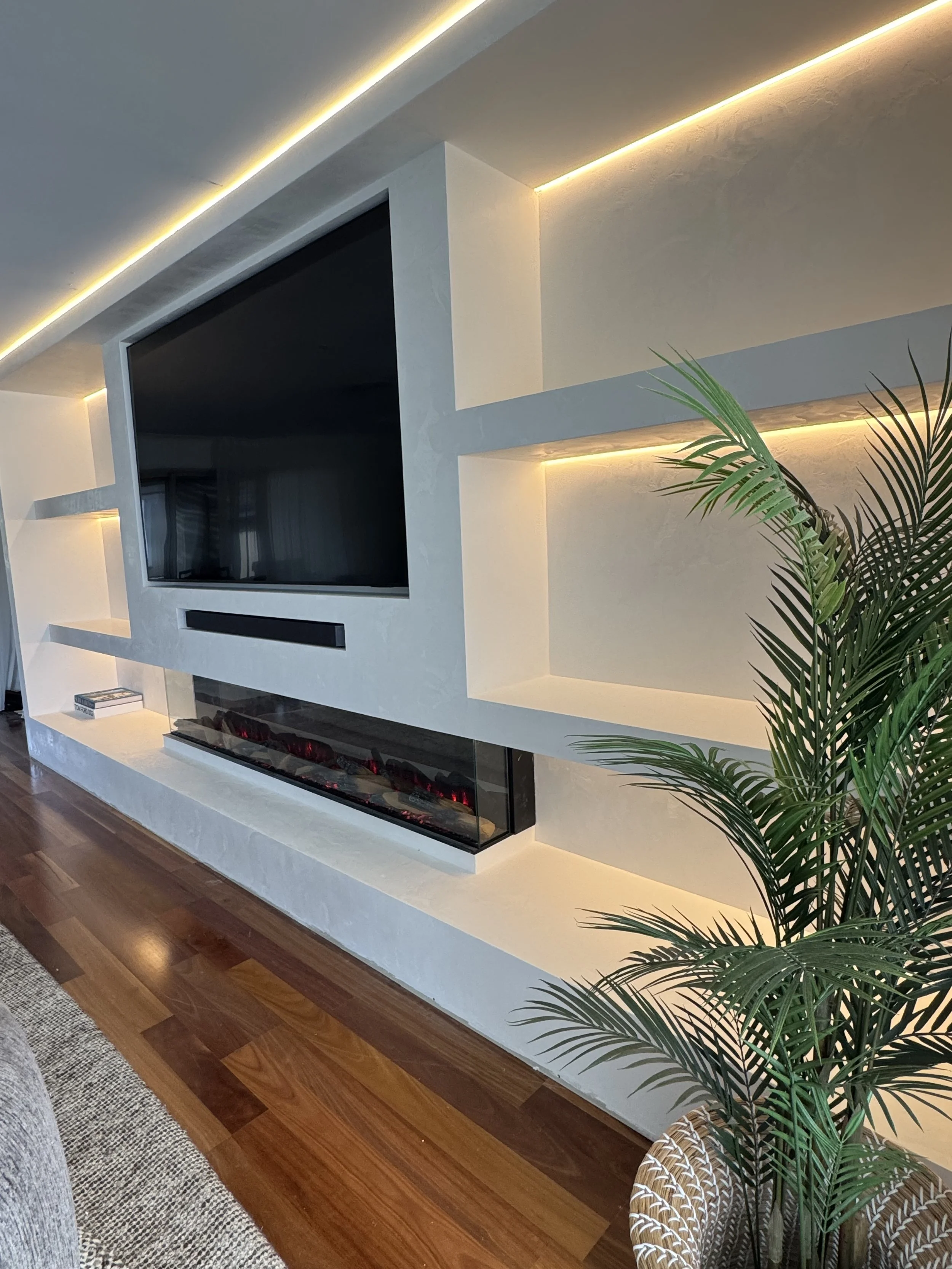 Modern living room with a wall-mounted flat screen TV, built-in shelving, illuminated ceiling lighting, a fireplace with red accents, hardwood floor, a gray sofa, a patterned armchair, and green indoor plants.