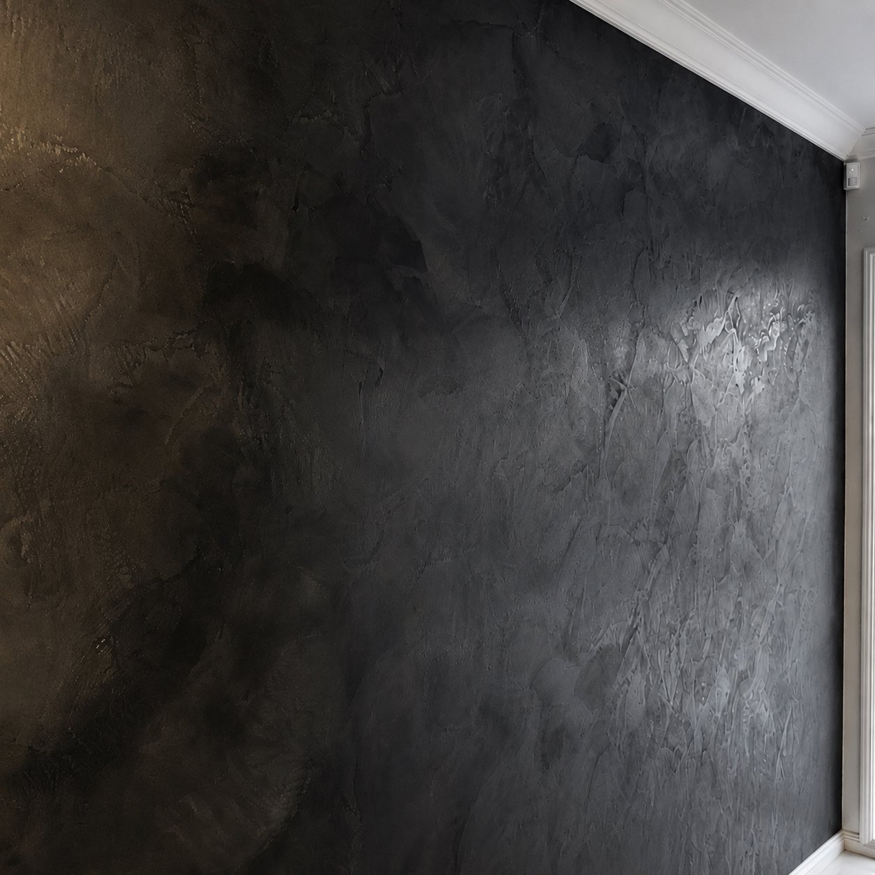 A dark, textured wall with a matte finish and a subtle wavy pattern, bordered by white crown molding and baseboard in a room with natural light.