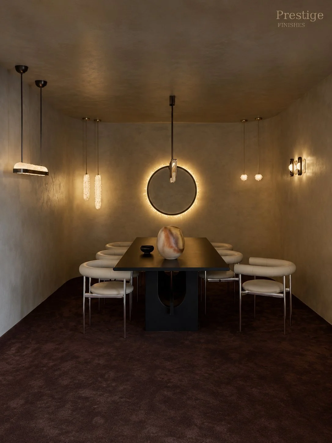 Modern dining room with six white chairs around a dark rectangular table, three decorative pendant lights, a circular mirror with backlighting on the wall, and a large decorative vase on the table.