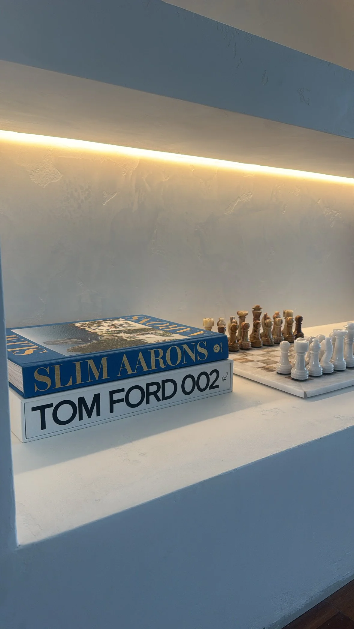 A white shelf with a stack of two books, one titled "SLIM AARONS" and the other "TOM FORD 002", alongside a chess set with white and brown chess pieces on a small chessboard.