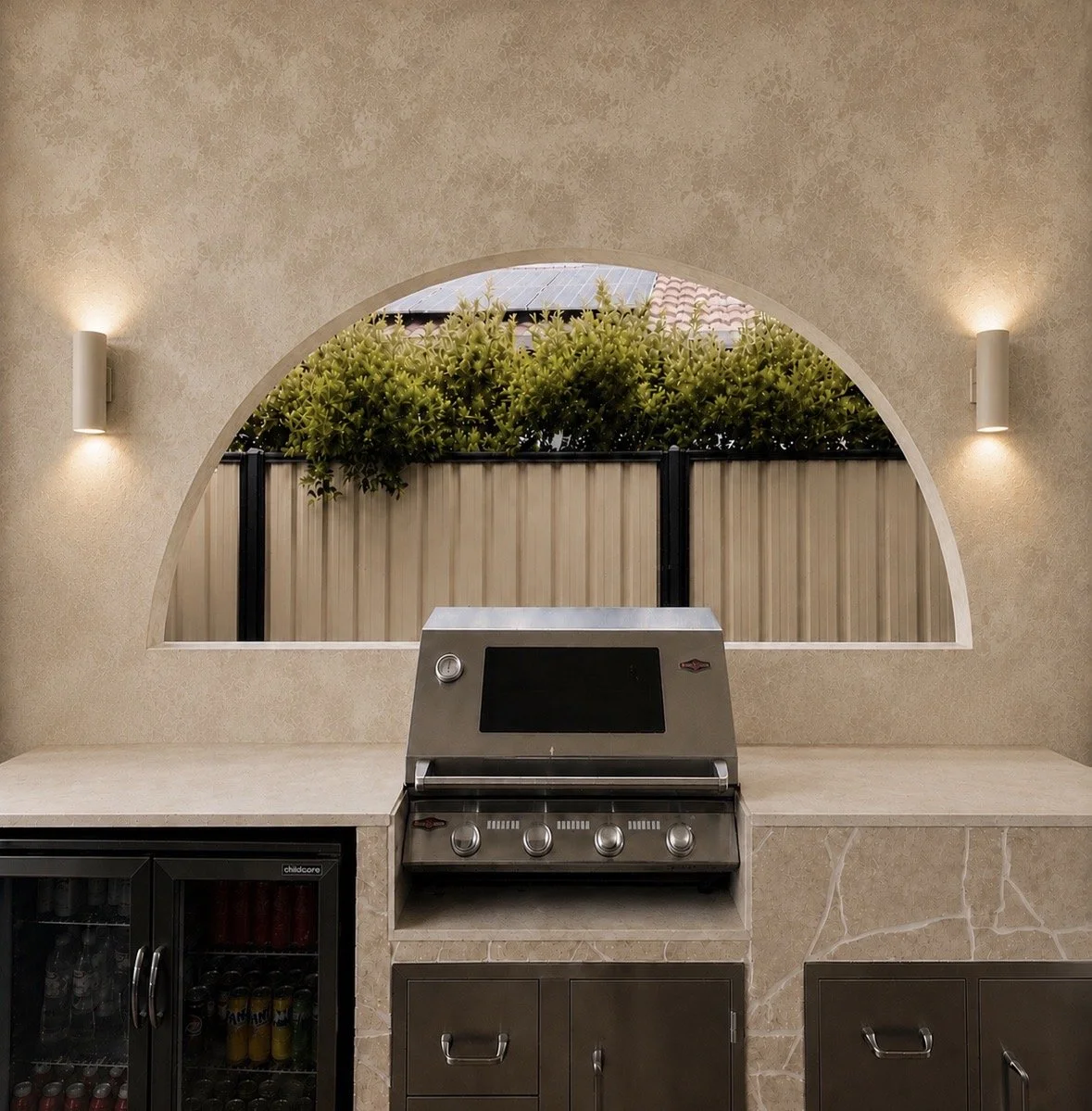 Outdoor kitchen with a built-in stainless steel grill, a counter, a wine cooler, and a large arched window showing green shrubbery and fencing.