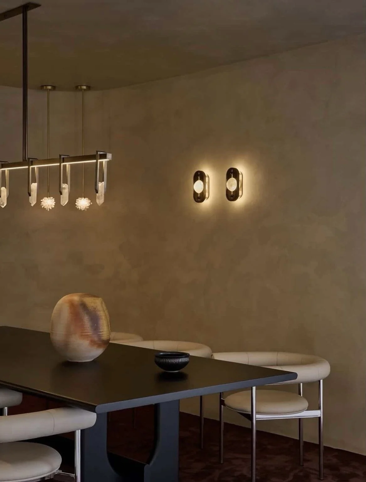 Modern dining room with a black table, cream-colored chairs, a decorative vase, and unique lighting fixtures on a beige wall.