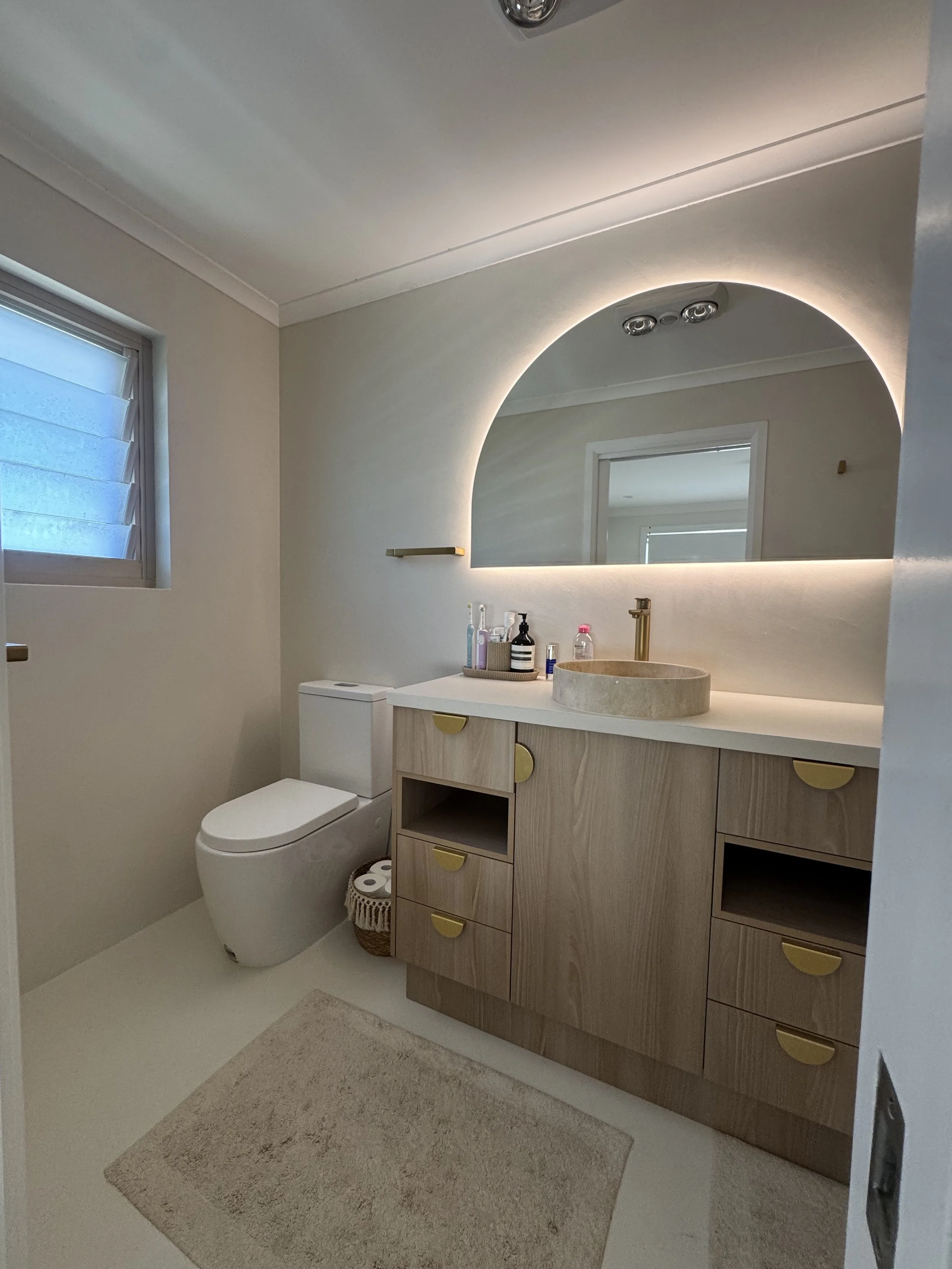 A modern bathroom with a large mirror, a beige vanity with storage drawers, a beige sink, and a toilet. There are toiletries on the vanity, a small wicker basket with toilet paper underneath the toilet, a window with blinds, and plush beige bath mats