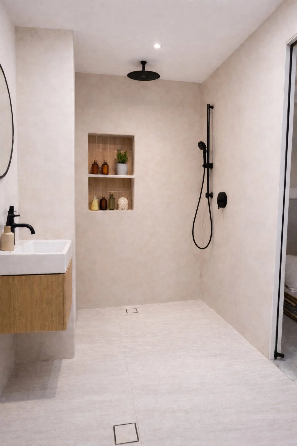 Minimalist bathroom with open shower area featuring black fixtures and a built-in niche with decorative bottles and a plant, a small white sink with a wooden cabinet, and a sliding door to an adjacent room.