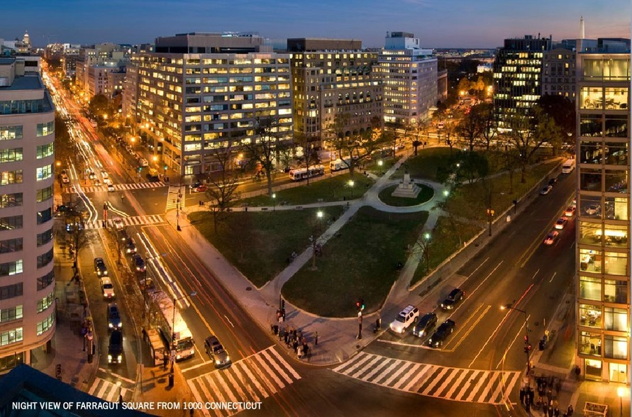 Night view of Farragut Square from 1717 K Street, NW office building in Washington, D.C., showing illuminated buildings, busy streets with moving cars, crosswalks, and a park with pathways and a monument in the center.
