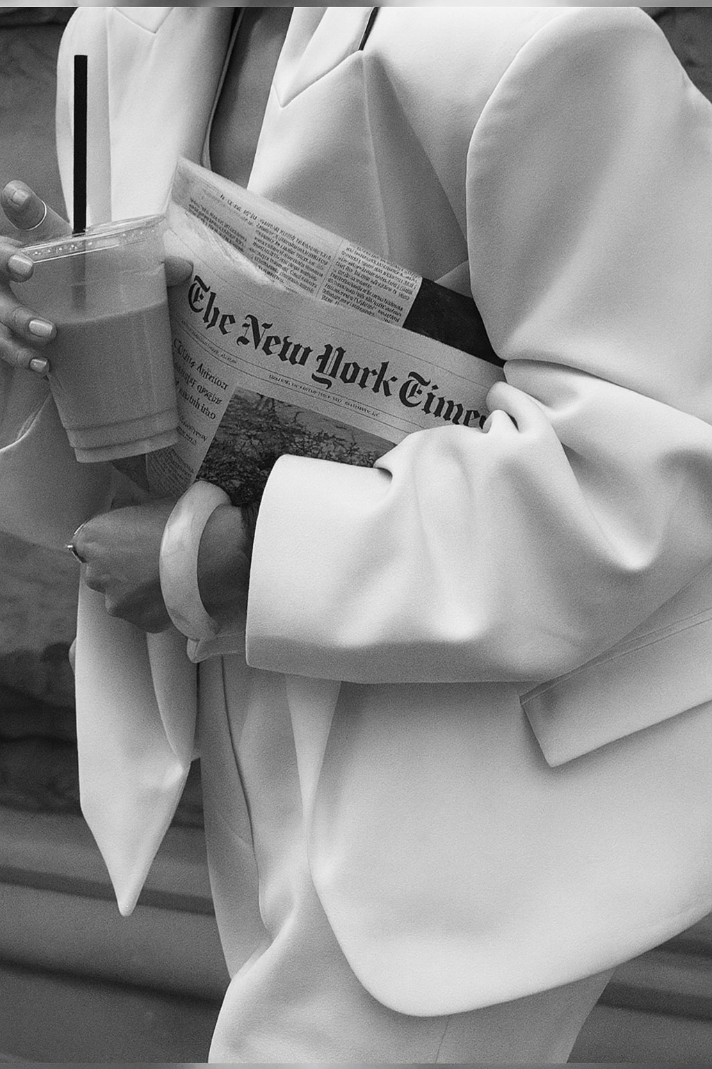Person holding a takeaway drink and a newspaper, with part of their torso and arm visible, wearing a white jacket and a bracelet.