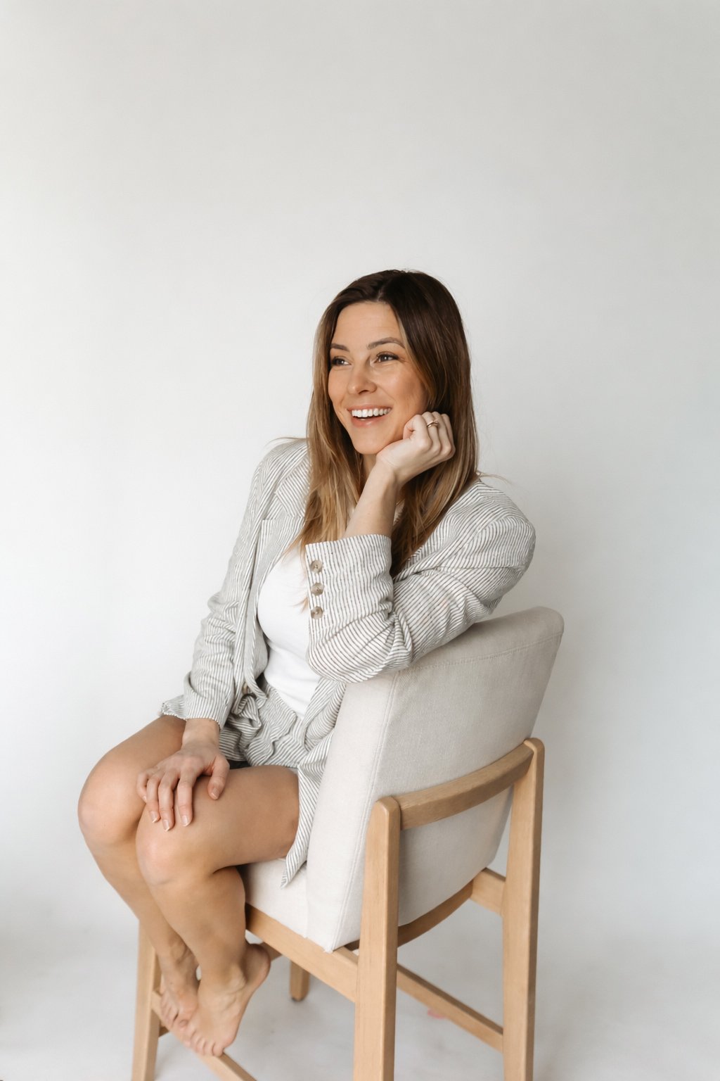 Woman sitting on a chair with a smile, wearing a light-colored blazer and white top, against a plain white background.