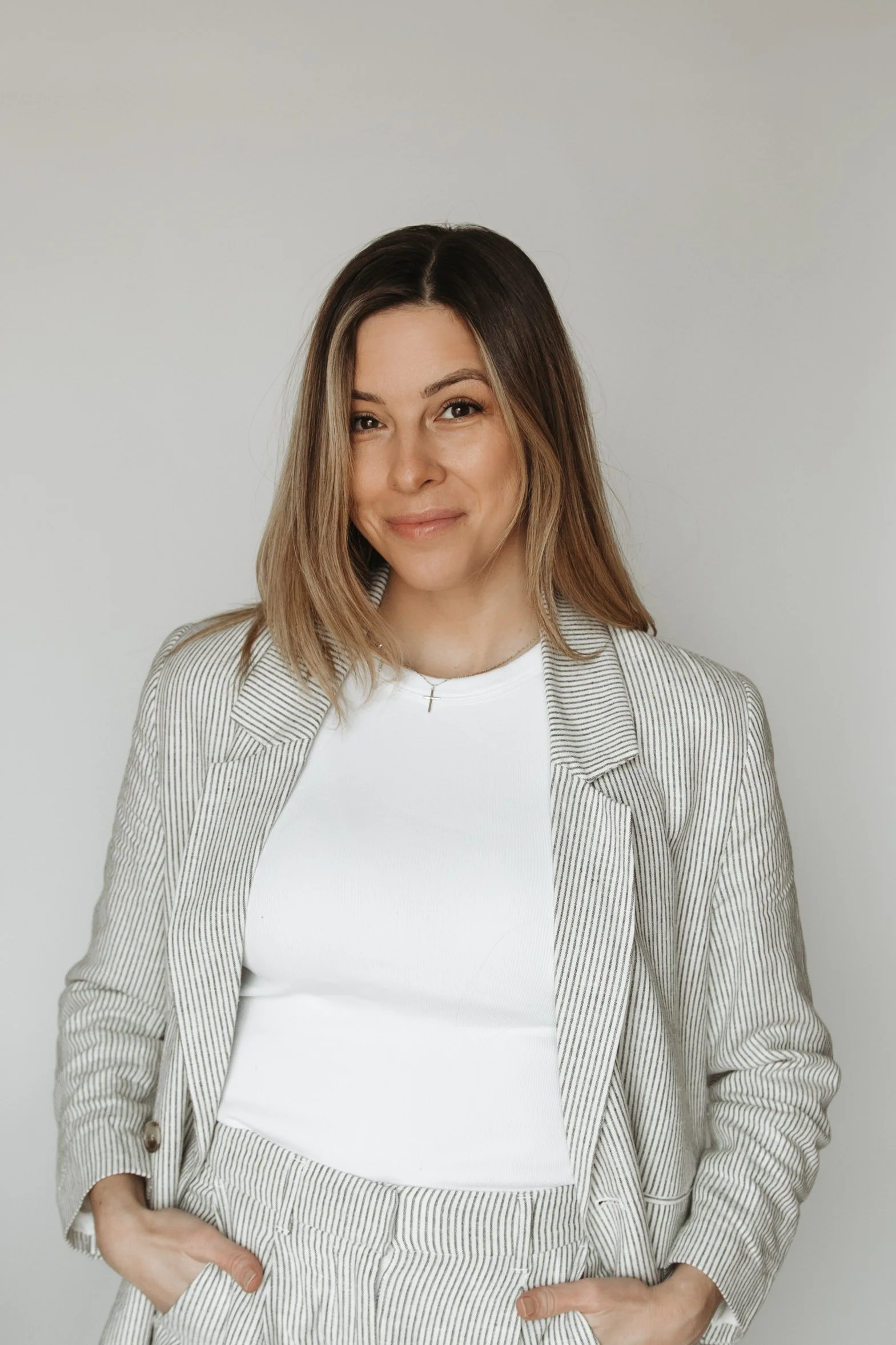 A woman with shoulder-length brown hair wearing a striped blazer and white t-shirt, standing against a plain gray background.