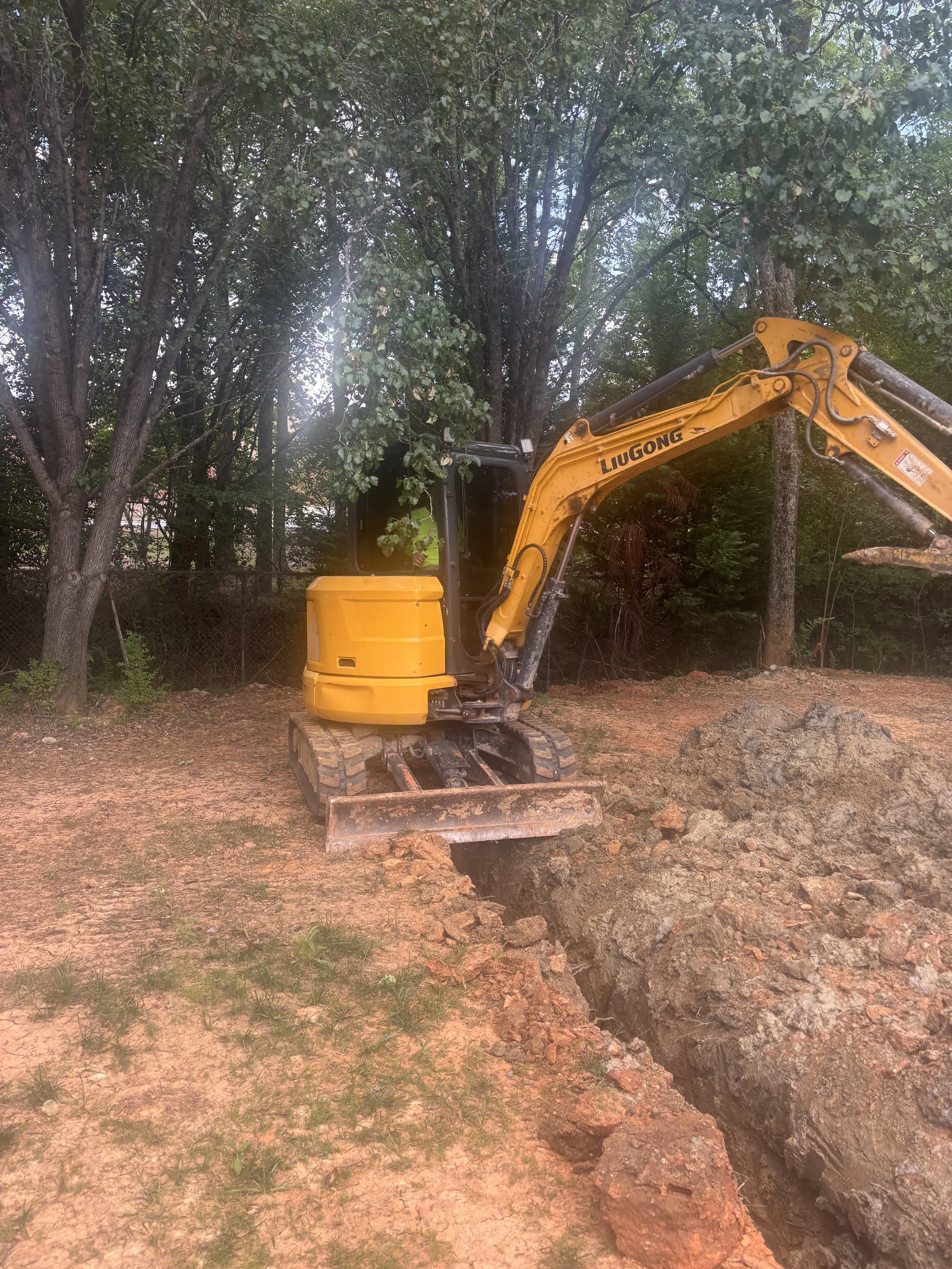 A yellow excavator digging a large hole in the ground in a wooded area.