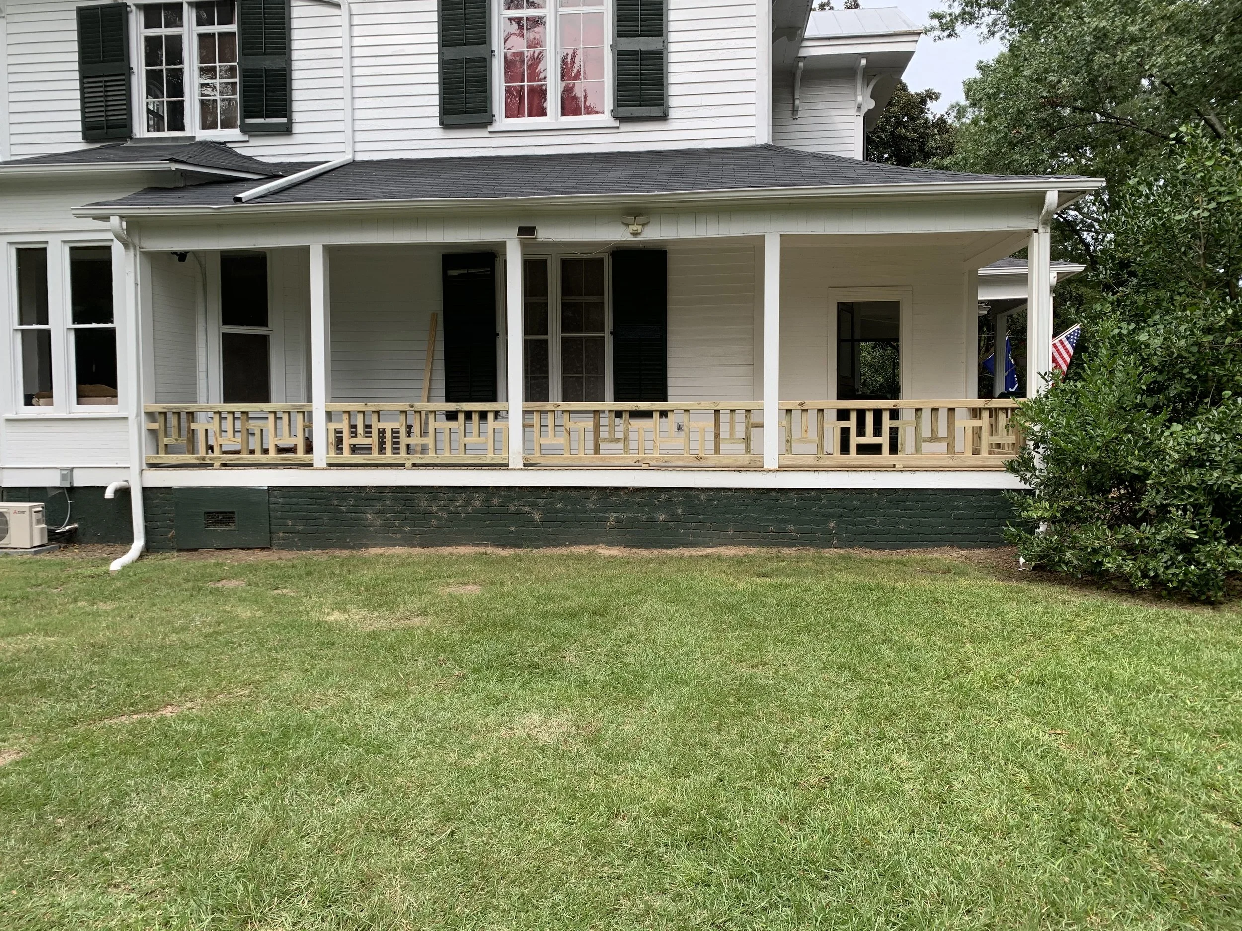 The image shows the back of a white, two-story house with large windows, black shutters, and a covered porch. There is a grassy yard in the foreground, a tree to the right, and some American flags near the house.