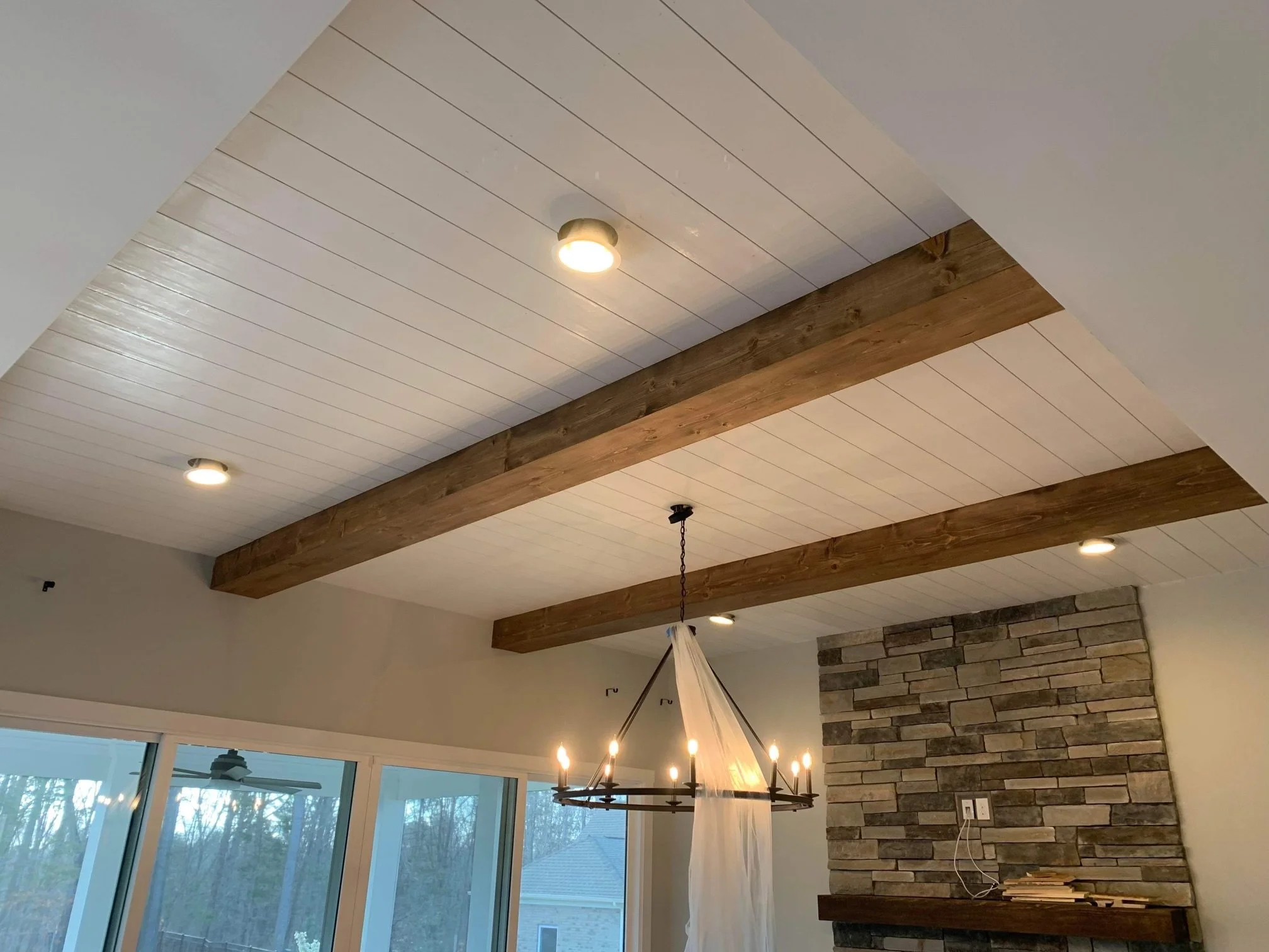 Ceiling with white wooden panels, exposed wooden beams, recessed lighting, a hanging chandelier with candles, large windows showing outdoor trees.