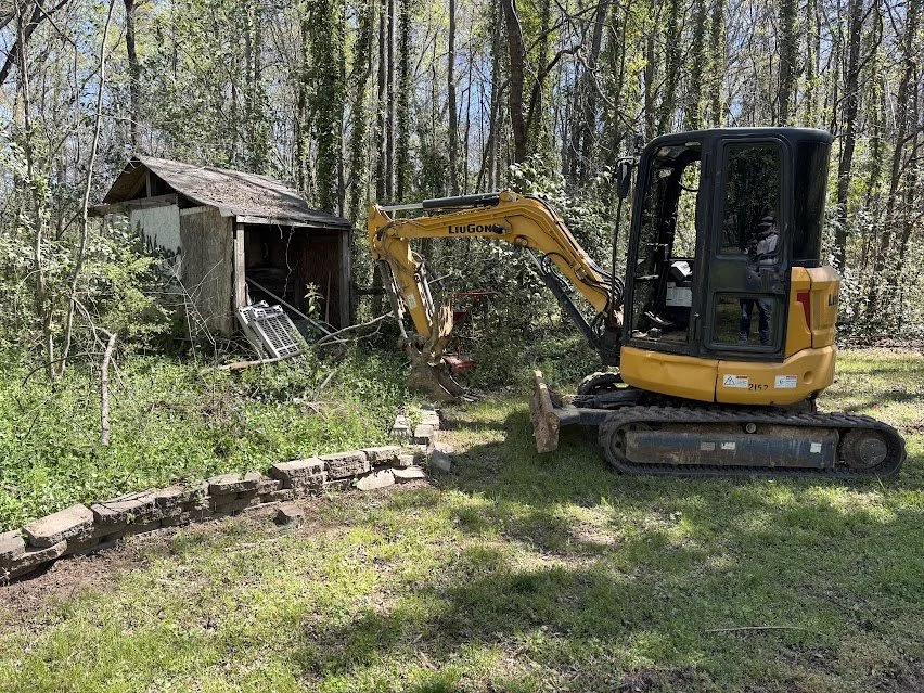 A yellow mini excavator clears debris near an old shed in a wooded yard.