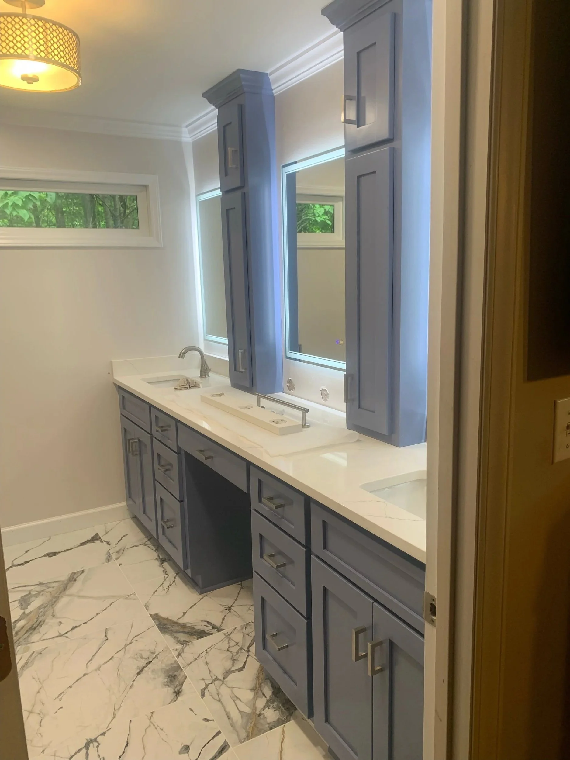Bathroom under construction with blue cabinetry, a white marble countertop, and marble floor tiles with gray veining. There is a window with greenery outside, and a light fixture on the ceiling.