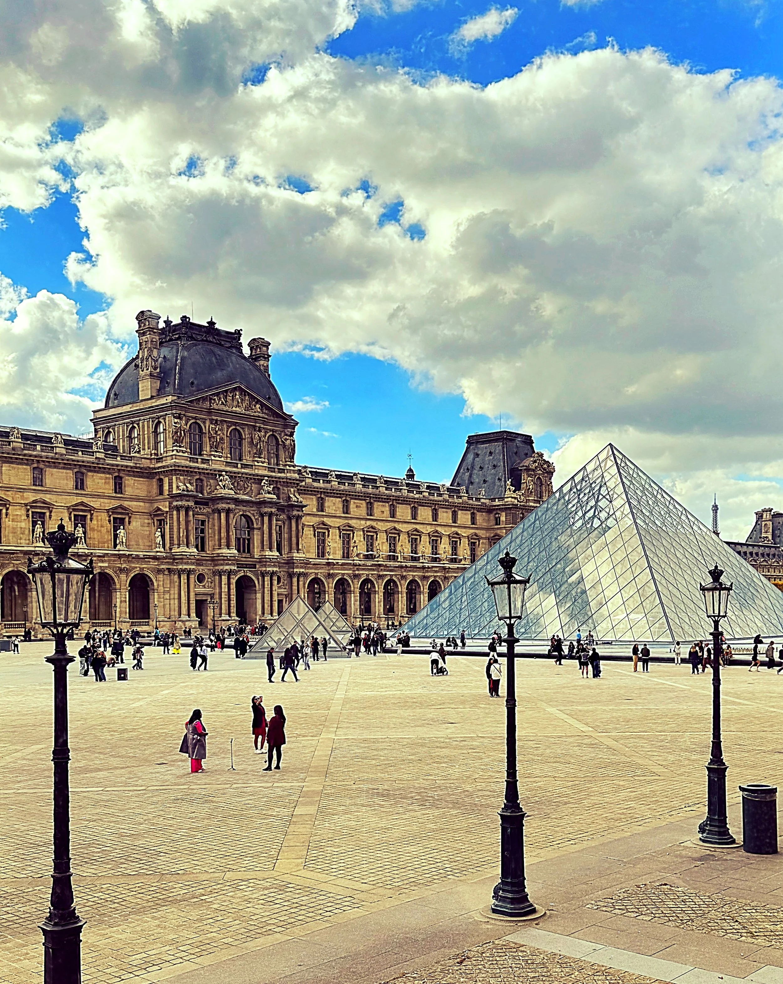 View of the Louvre Museum in Paris, France, with its glass pyramid entrance, historic architecture, and people walking on the courtyard under a partly cloudy sky.