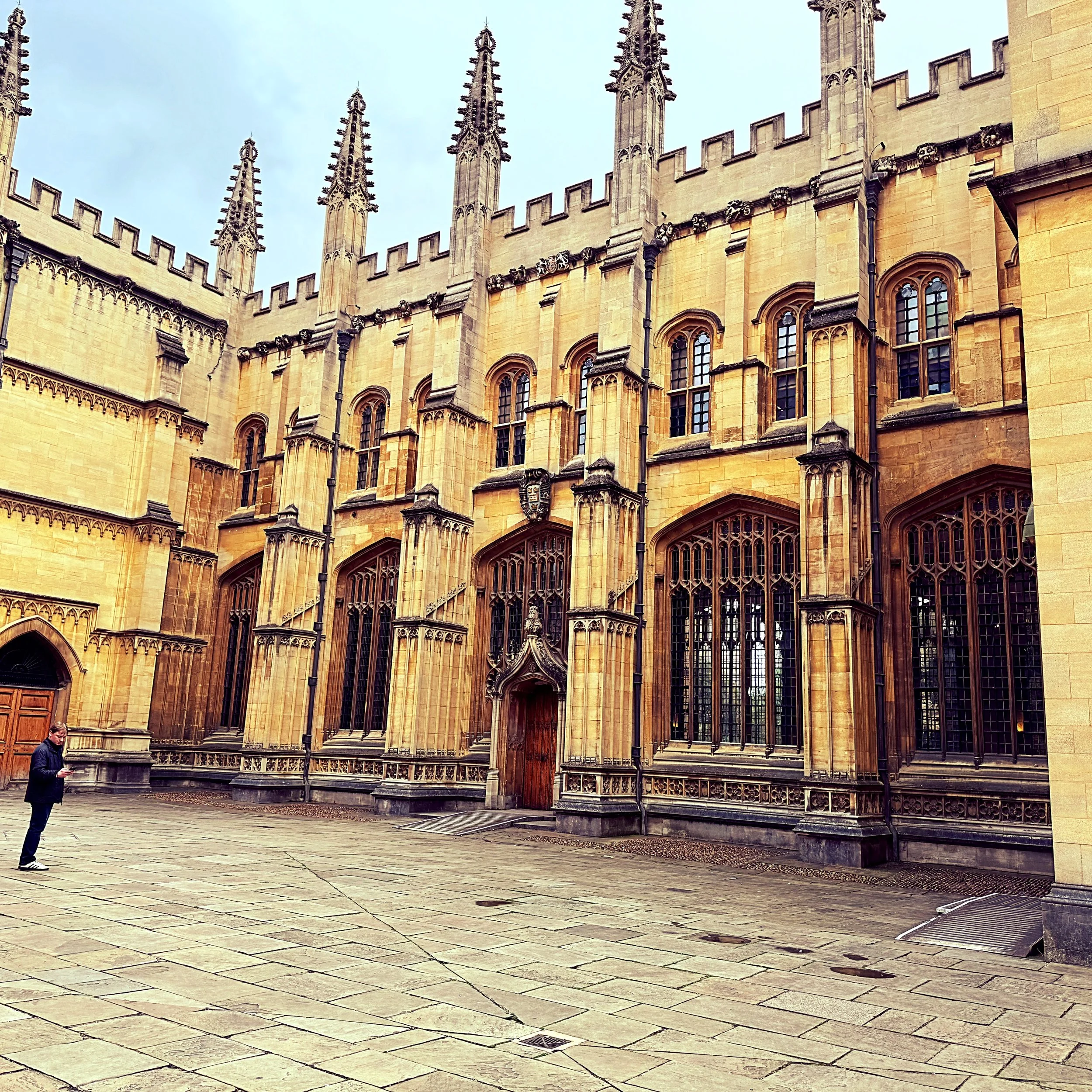 A historic stone building with gothic architectural features, including tall spires, arched windows, and decorative carvings, seen on a cloudy day. A man in a dark jacket and glasses is walking at the bottom left, looking at his phone.